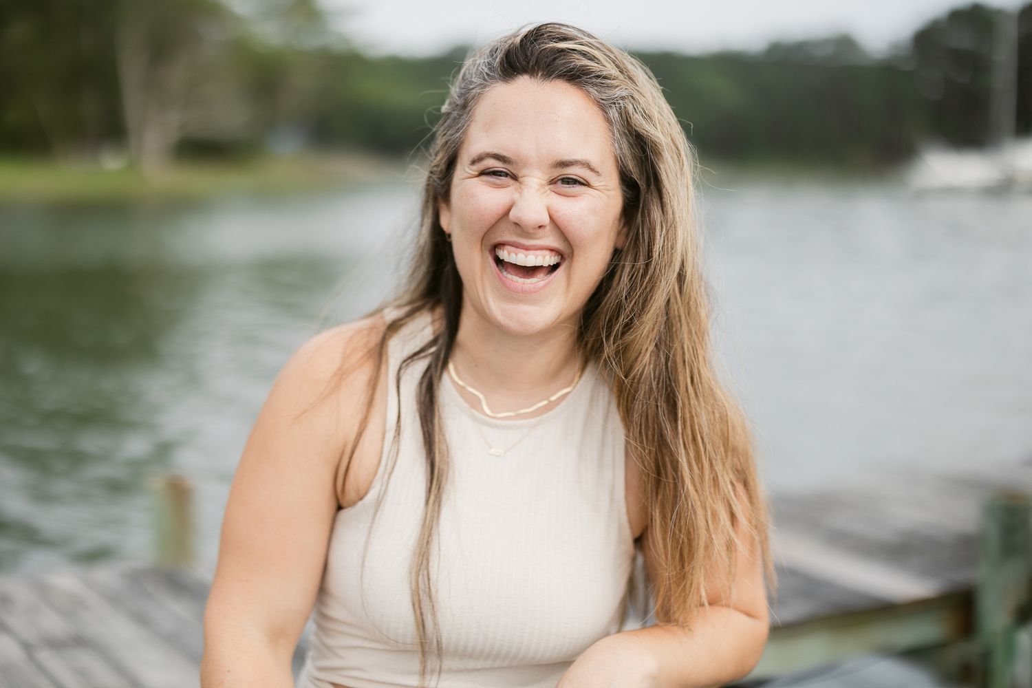 A joyful person with long hair wearing a cream tank top laughs heartily near a lake on a summer day.