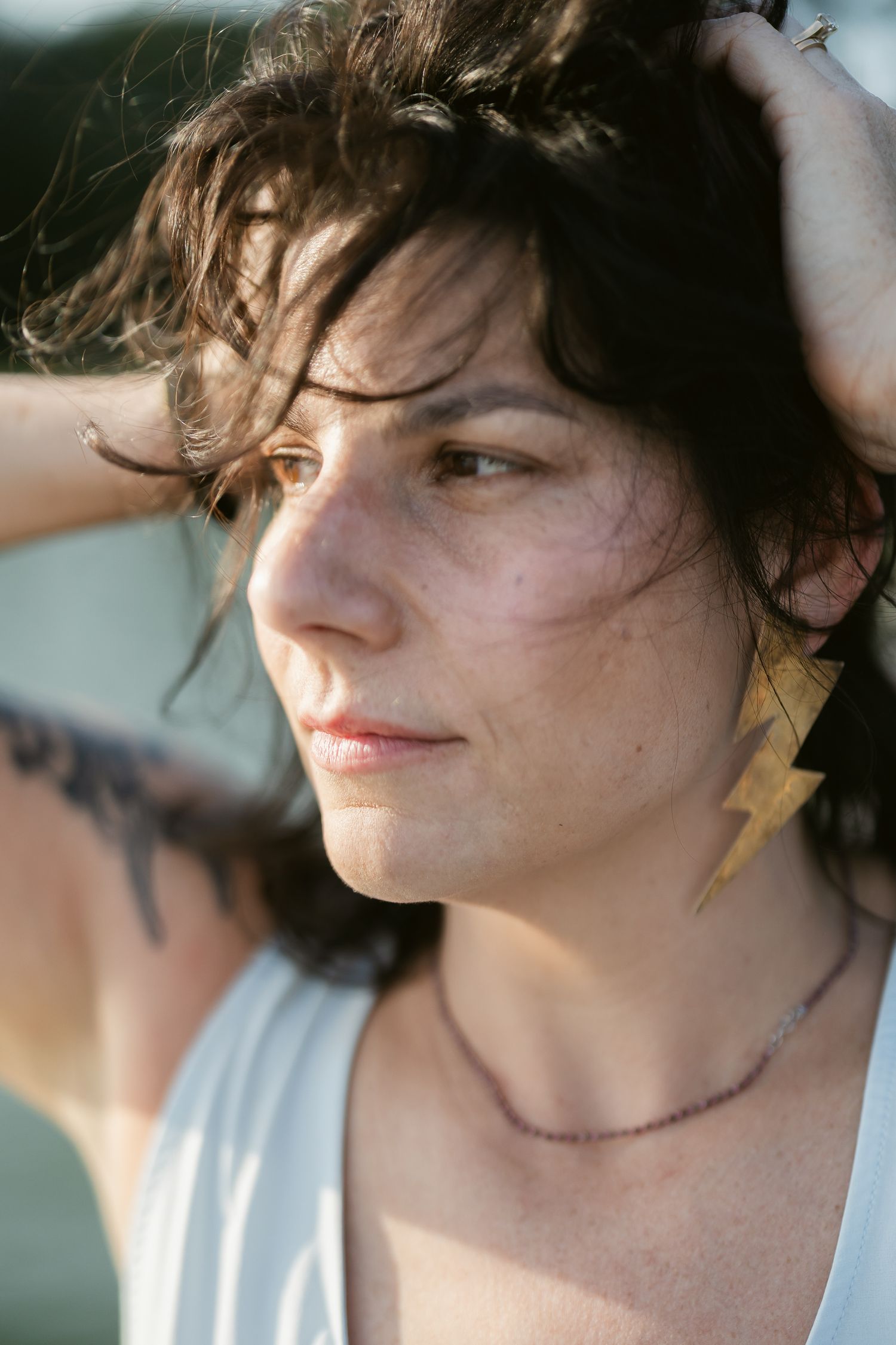 Person in white tank top with tousled dark hair looking pensively to the side in natural lighting.