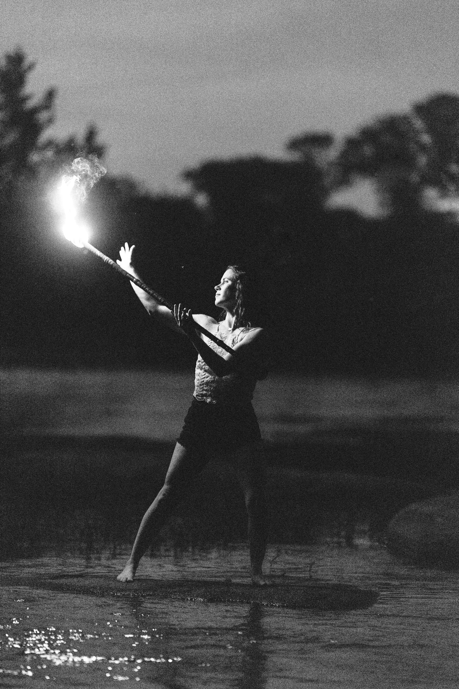 A dramatic black and white photo of a person holding a light while standing in shallow water at dusk.