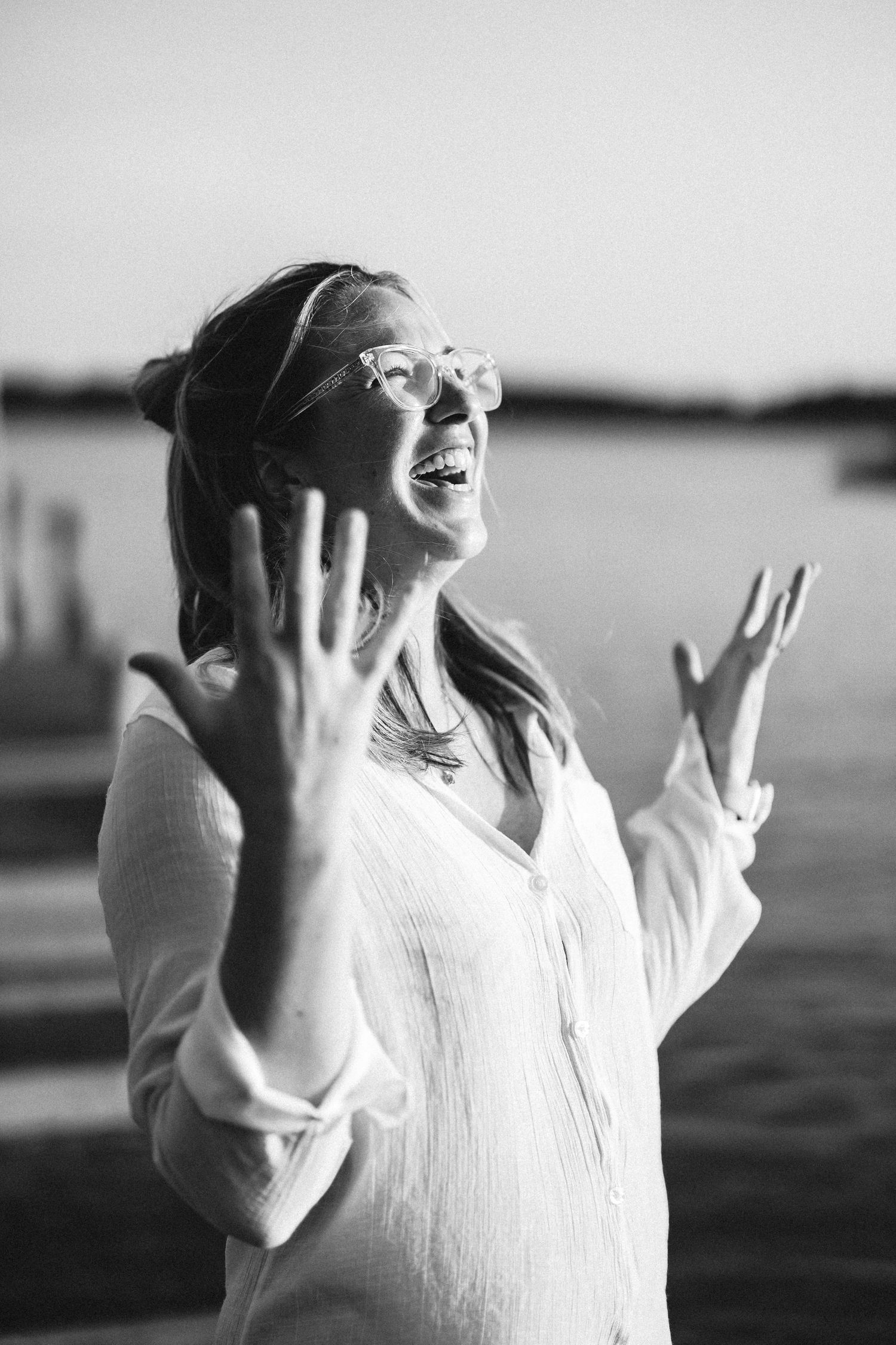 A person in white clothing raises their arms joyfully against a blurred outdoor background in black and white.