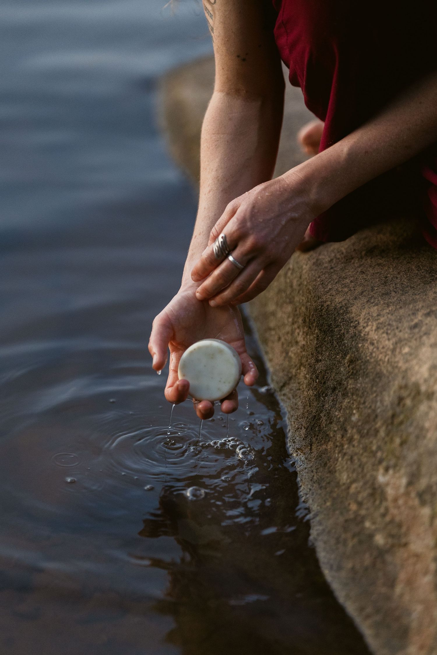 Hands gently touch water while holding a smooth stone at the edge of a lake in warm sunset lighting.