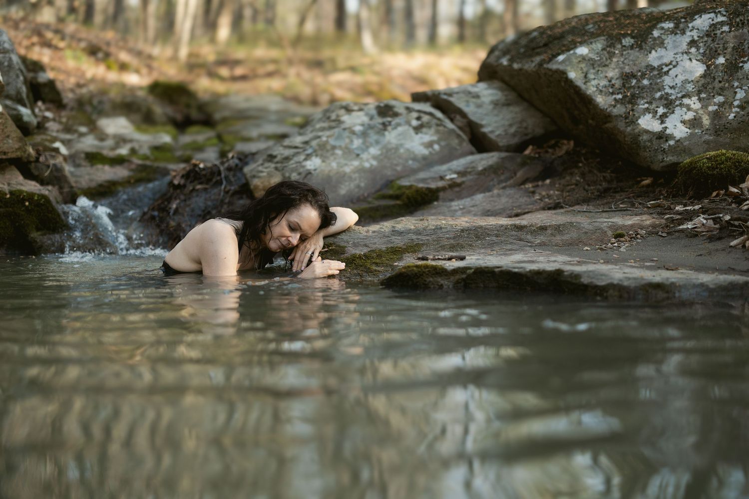 A person relaxes in a serene natural pool surrounded by rocks in a forest setting.