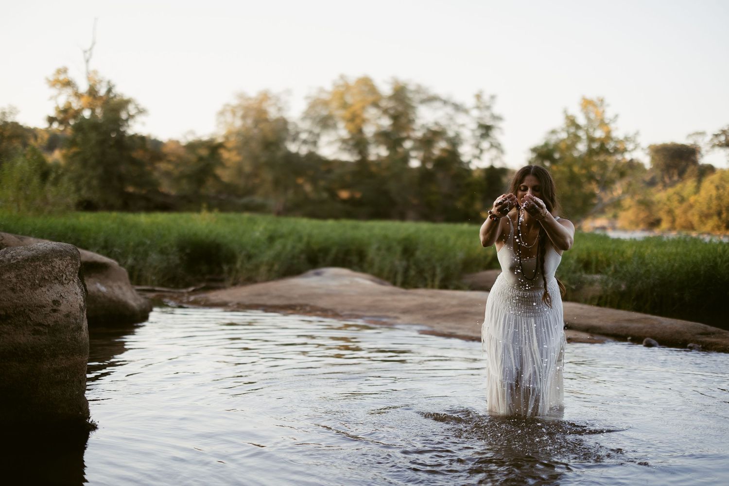 Person wearing white dress stands in flowing river water during golden sunset hour creating ethereal atmosphere.
