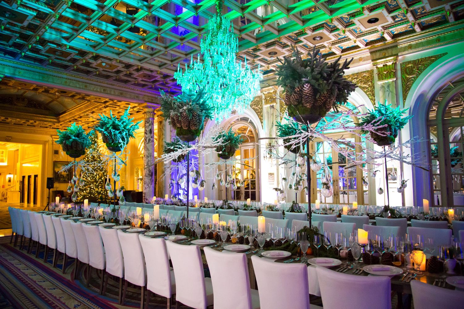 Elegant banquet table setting with teal and purple lighting beneath ornate ceiling with hanging floral arrangements.