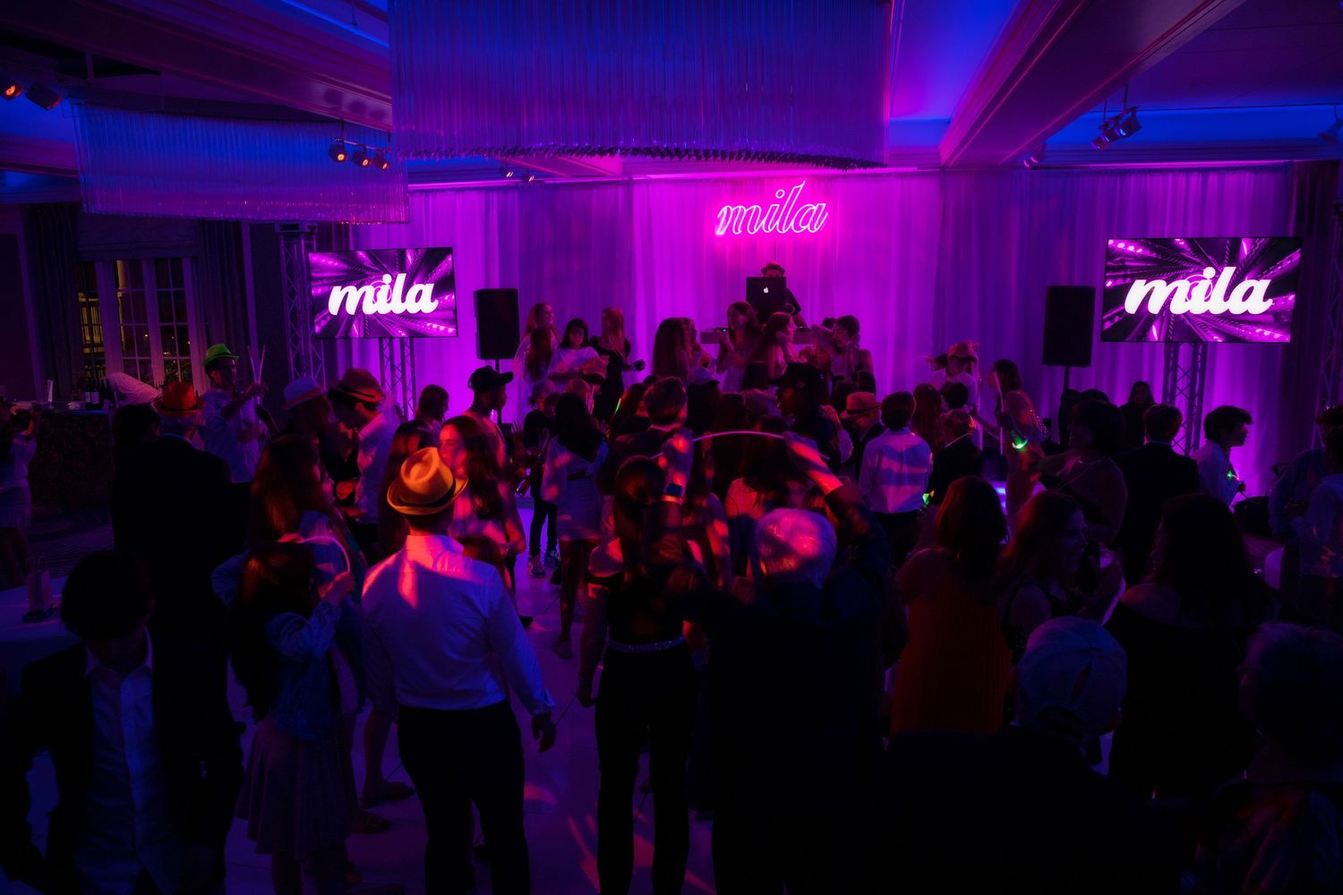 Crowded nightclub dance floor illuminated by pink neon 'mila' signs.