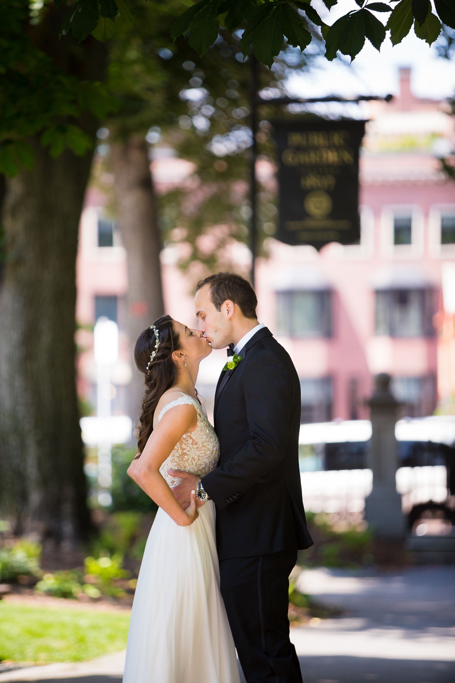Romantic wedding kiss in a park setting with sunlit trees and a vintage sign.