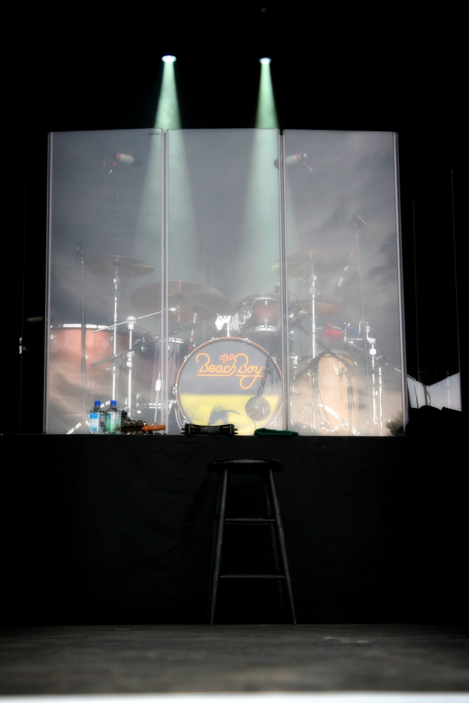 Drum kit silhouetted behind translucent screen with dramatic spotlight beams.