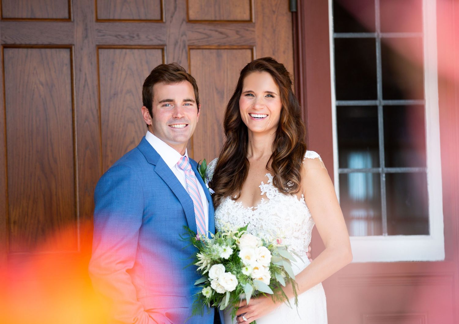 A couple in wedding attire pose together against a rustic wooden wall with warm lighting.