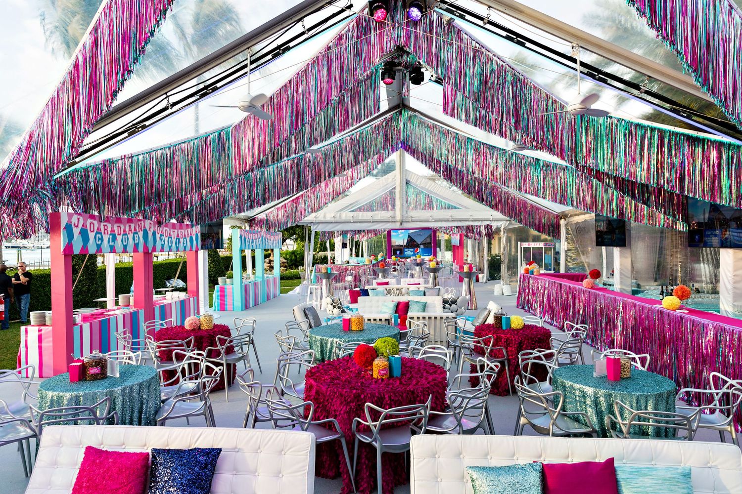 Pink and white streamers decorate a tent-covered outdoor event space with blue chairs and tables.