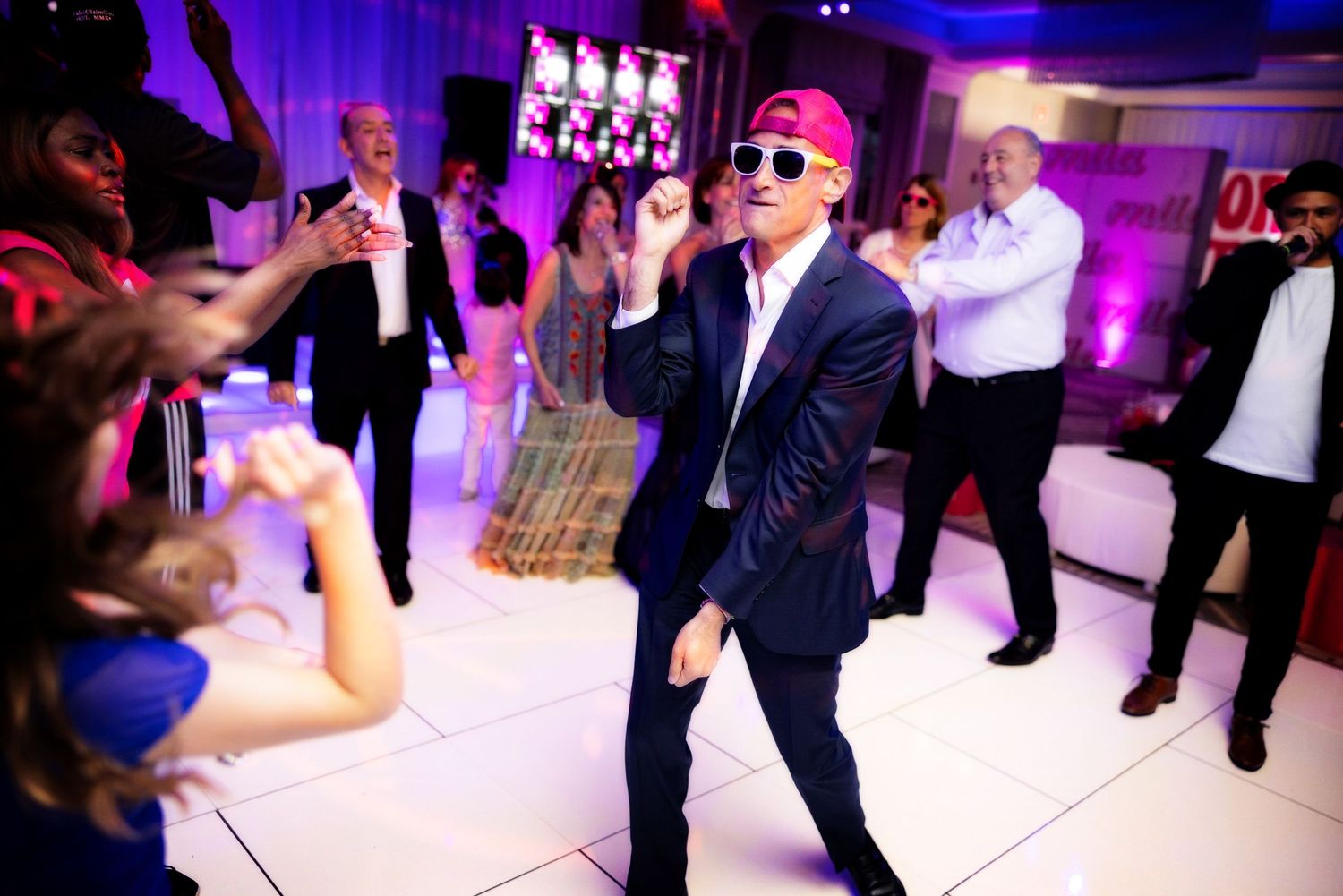 Party guest in sunglasses and suit dancing on illuminated dance floor surrounded by crowd.