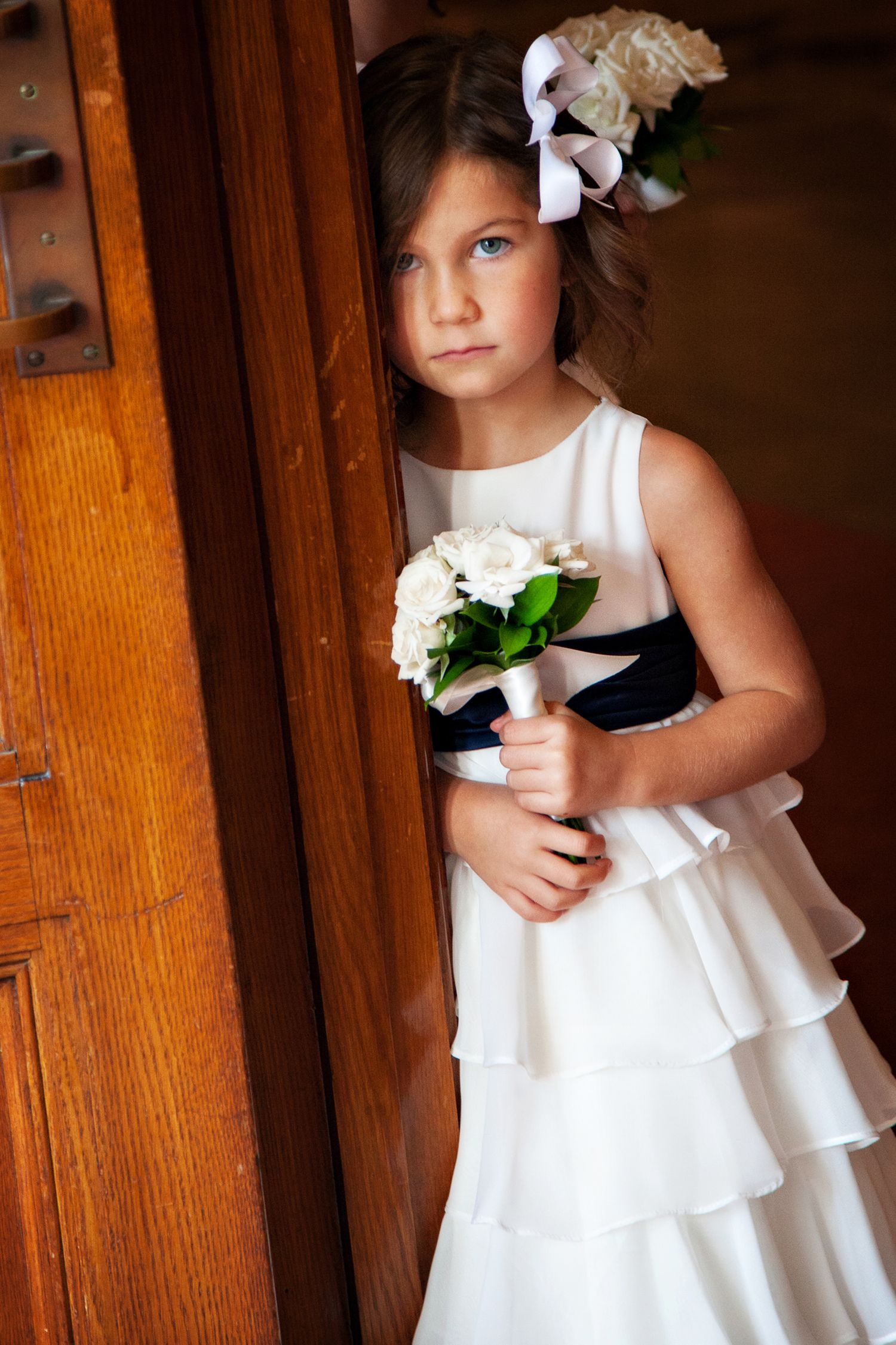 A flower girl in a white ruffled dress holds a small bouquet against a wooden door background.