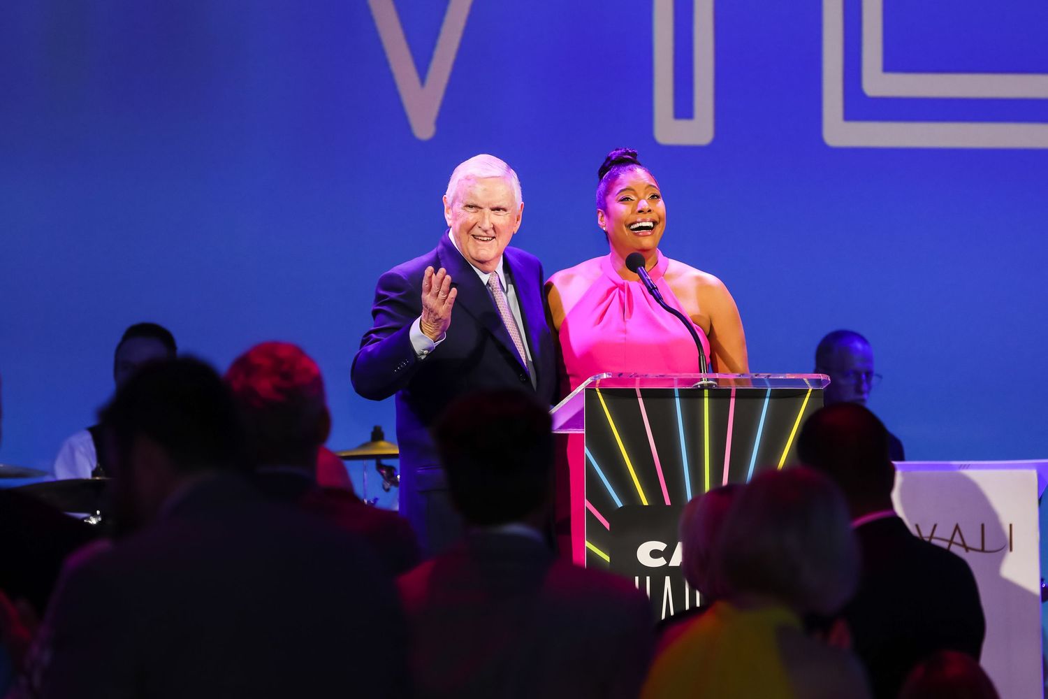 Two presenters speaking at a formal event on stage with blue backdrop.