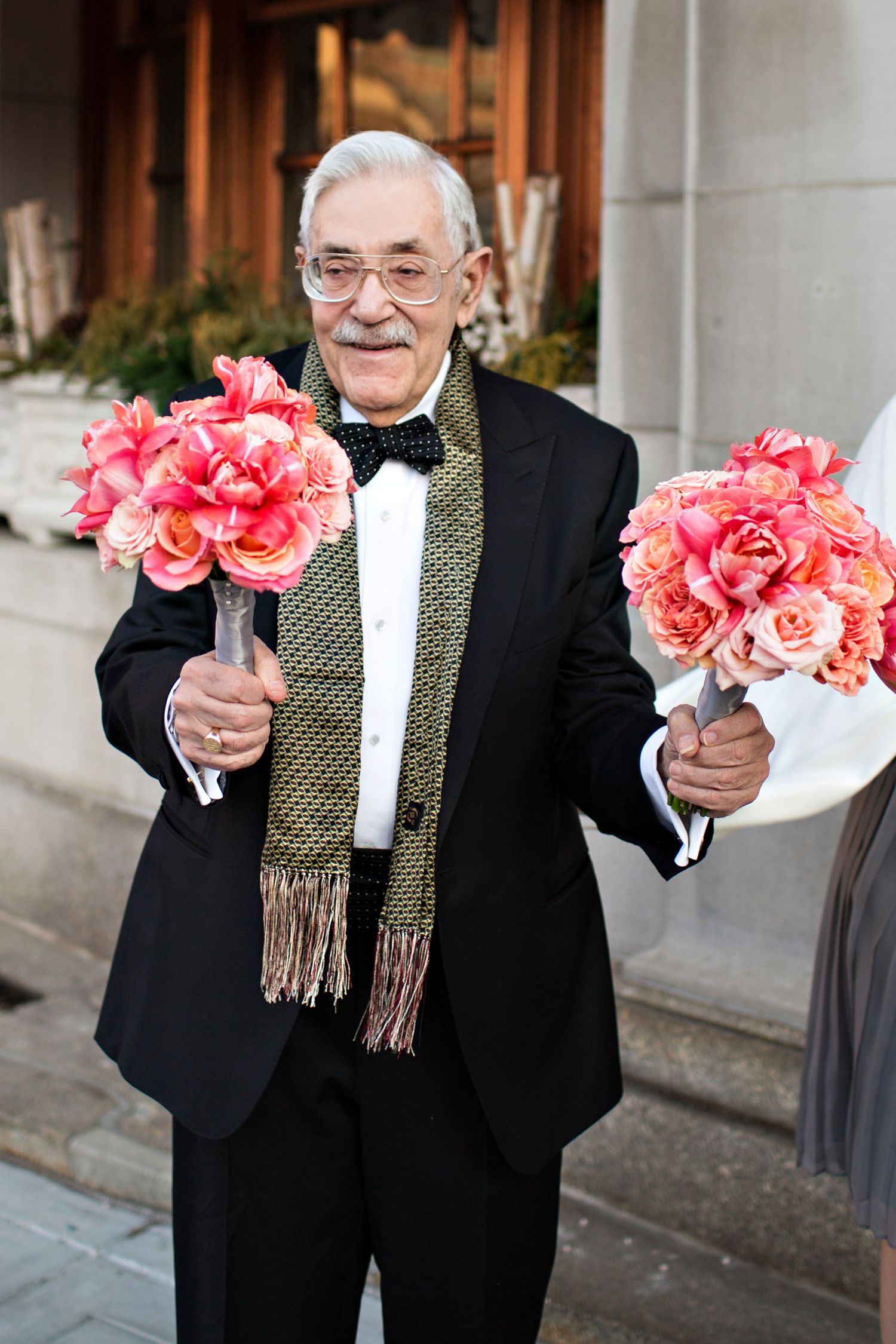 Person in formal black suit and scarf holding a bouquet of vibrant pink and coral roses against a stone building exterior.