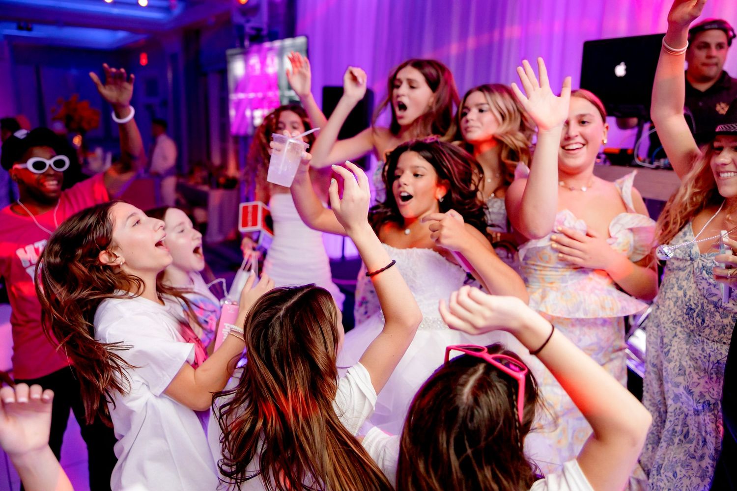 Enthusiastic party crowd dancing with hands in the air under purple lighting.