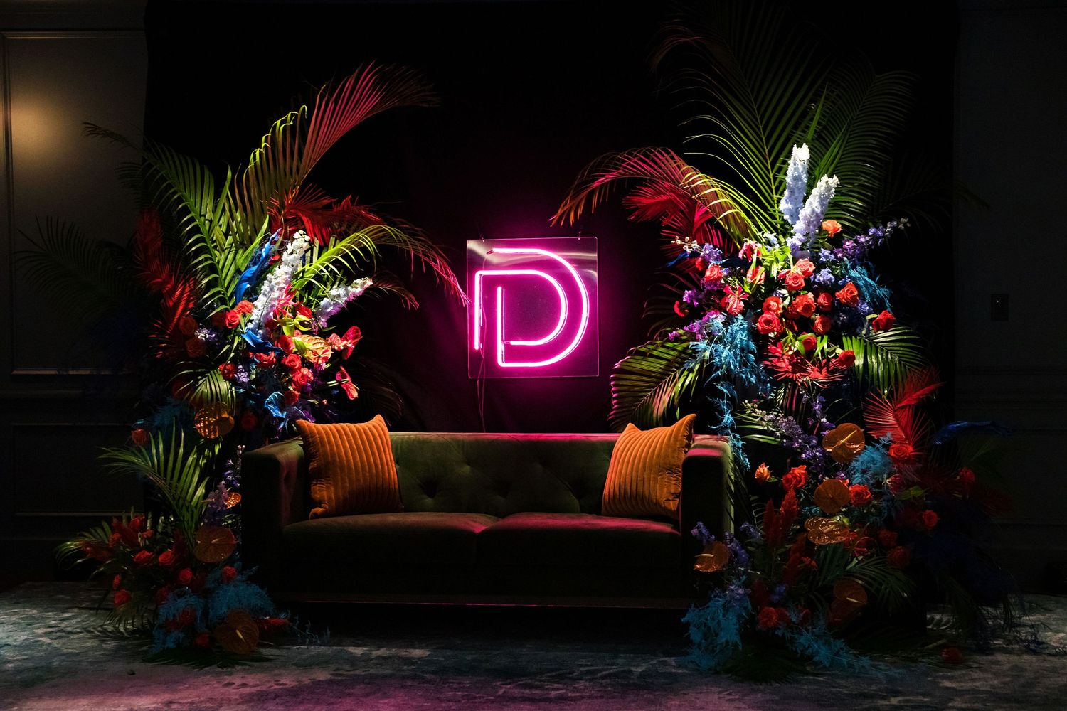Stylish lounge setting with pink neon D sign flanked by tropical floral arrangements and velvet couch.