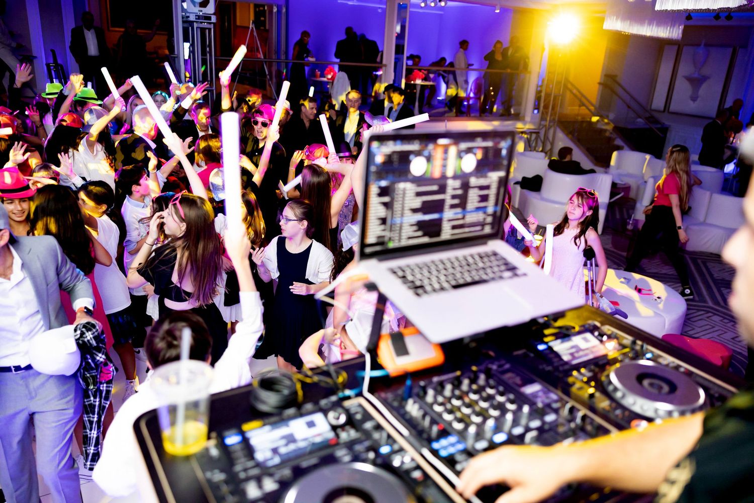 DJ setup with laptop and mixer while crowd dances in background under party lights.
