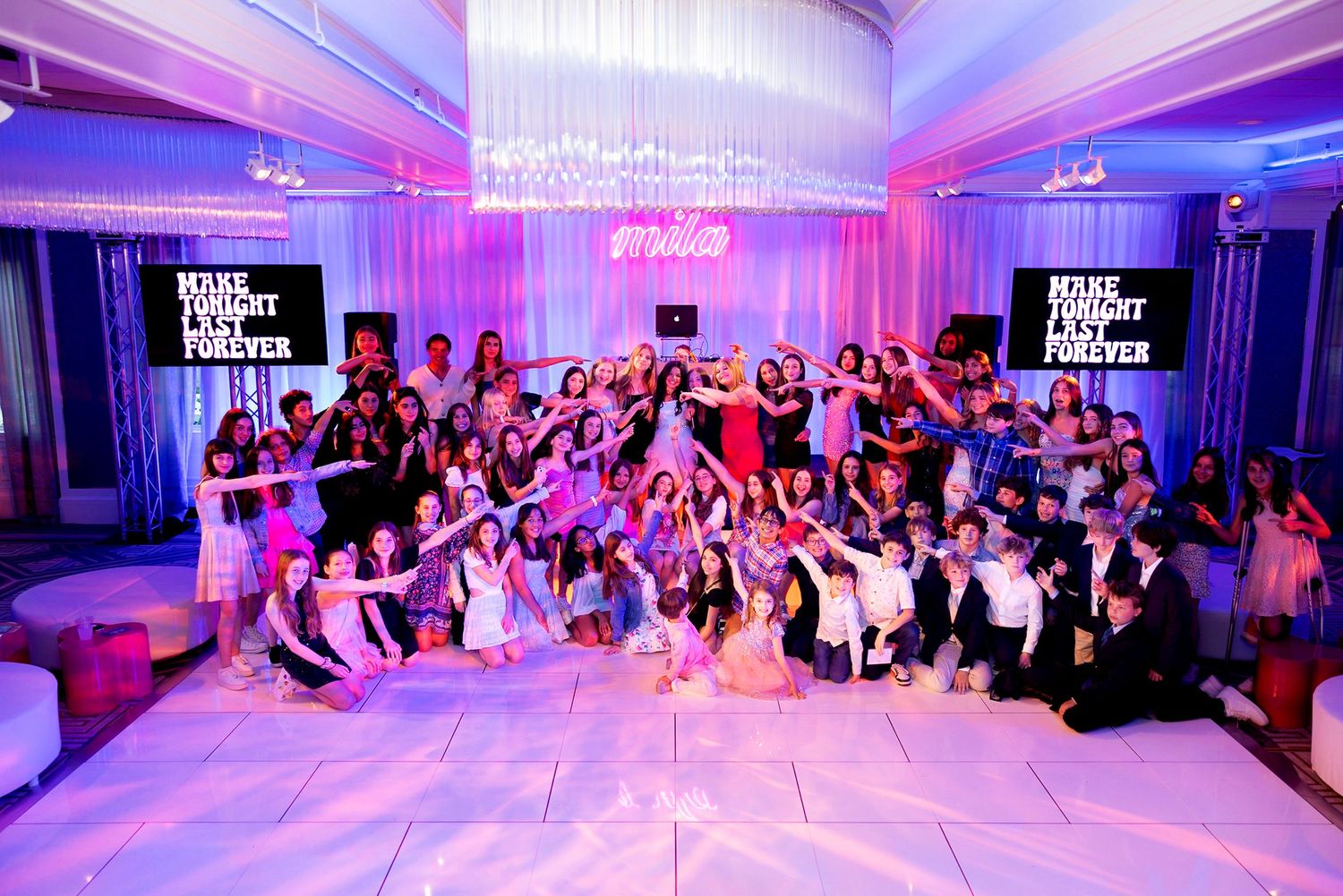 Large group photo of party attendees posing together in a decorated ballroom with pink lighting.