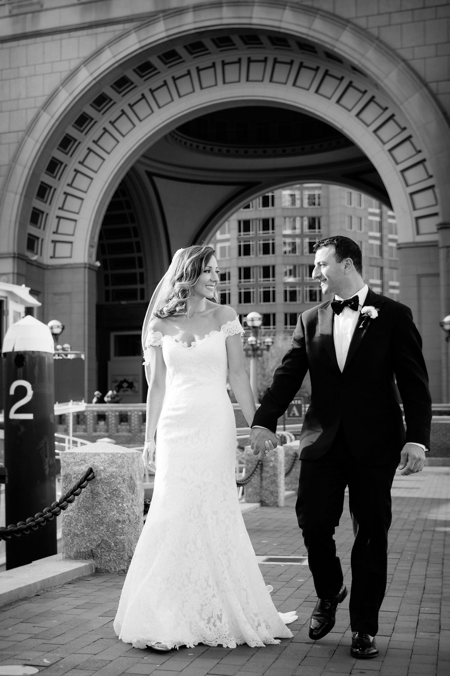 Elegant couple in wedding attire walking under ornate stone archway.