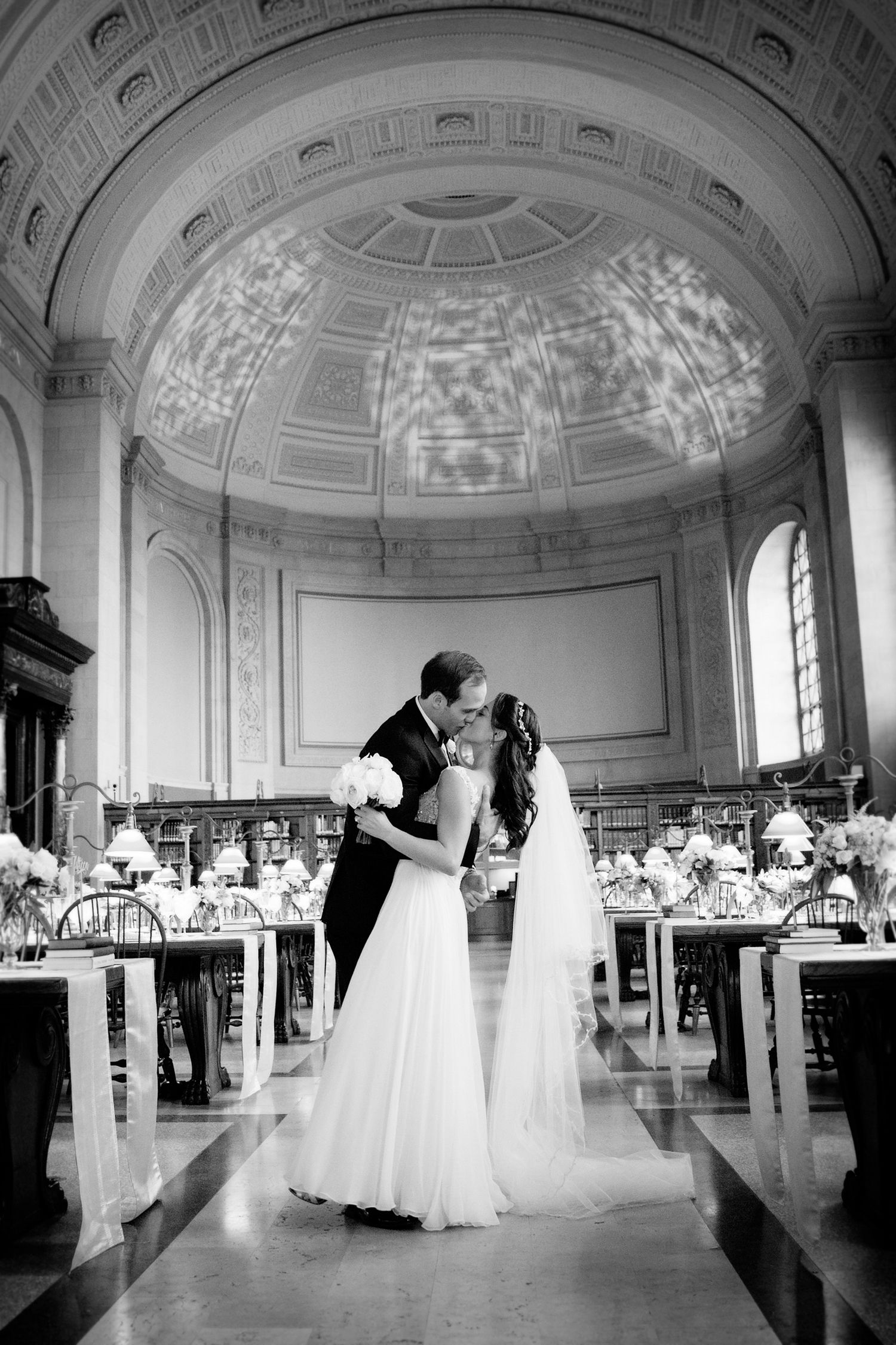 Wedding couple shares a kiss under an ornate domed ceiling in a grand ballroom.