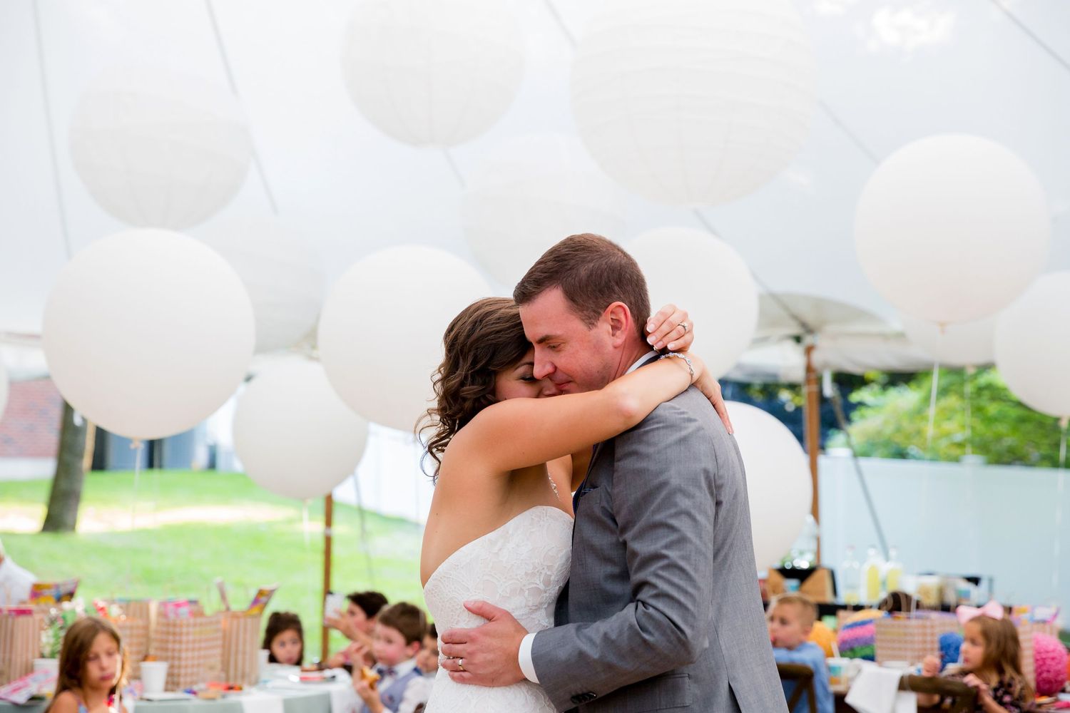 A couple shares a romantic first dance at their outdoor wedding reception surrounded by white balloons and guests.