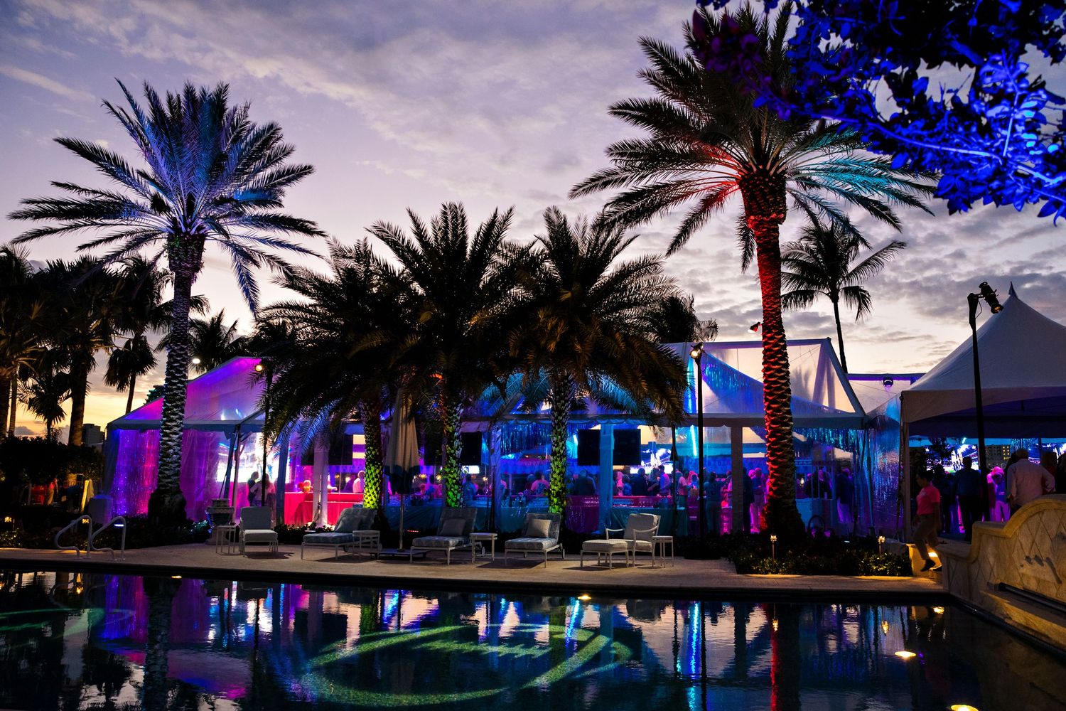 Palm trees silhouetted against twilight sky with colorful illuminated poolside venue.