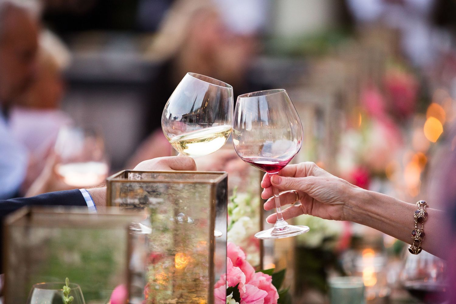 A romantic outdoor dining setup with candles, flowers and wine glasses on a rustic wooden table at sunset.