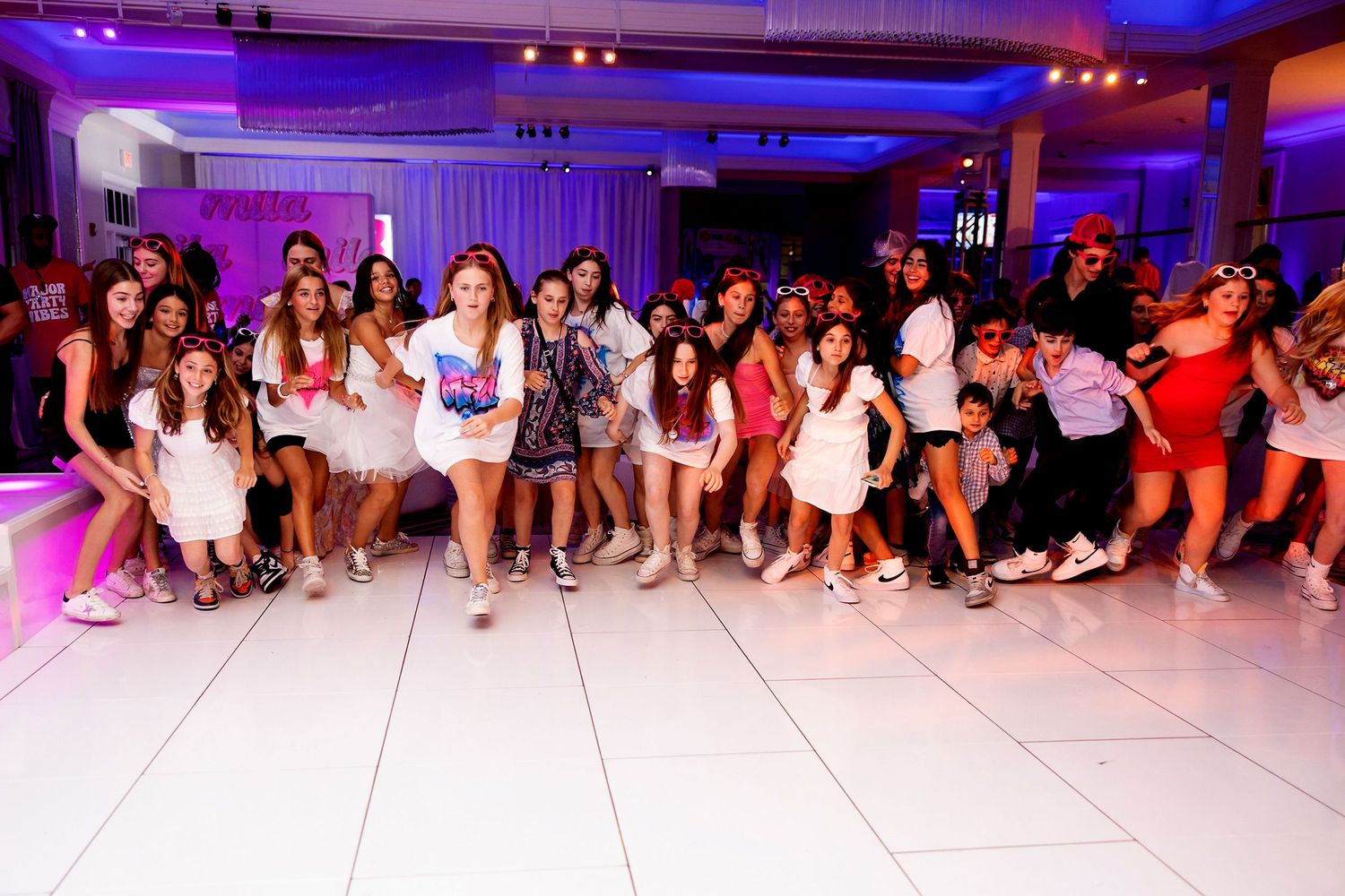 Large group of party guests posing together on white dance floor under pink lighting.