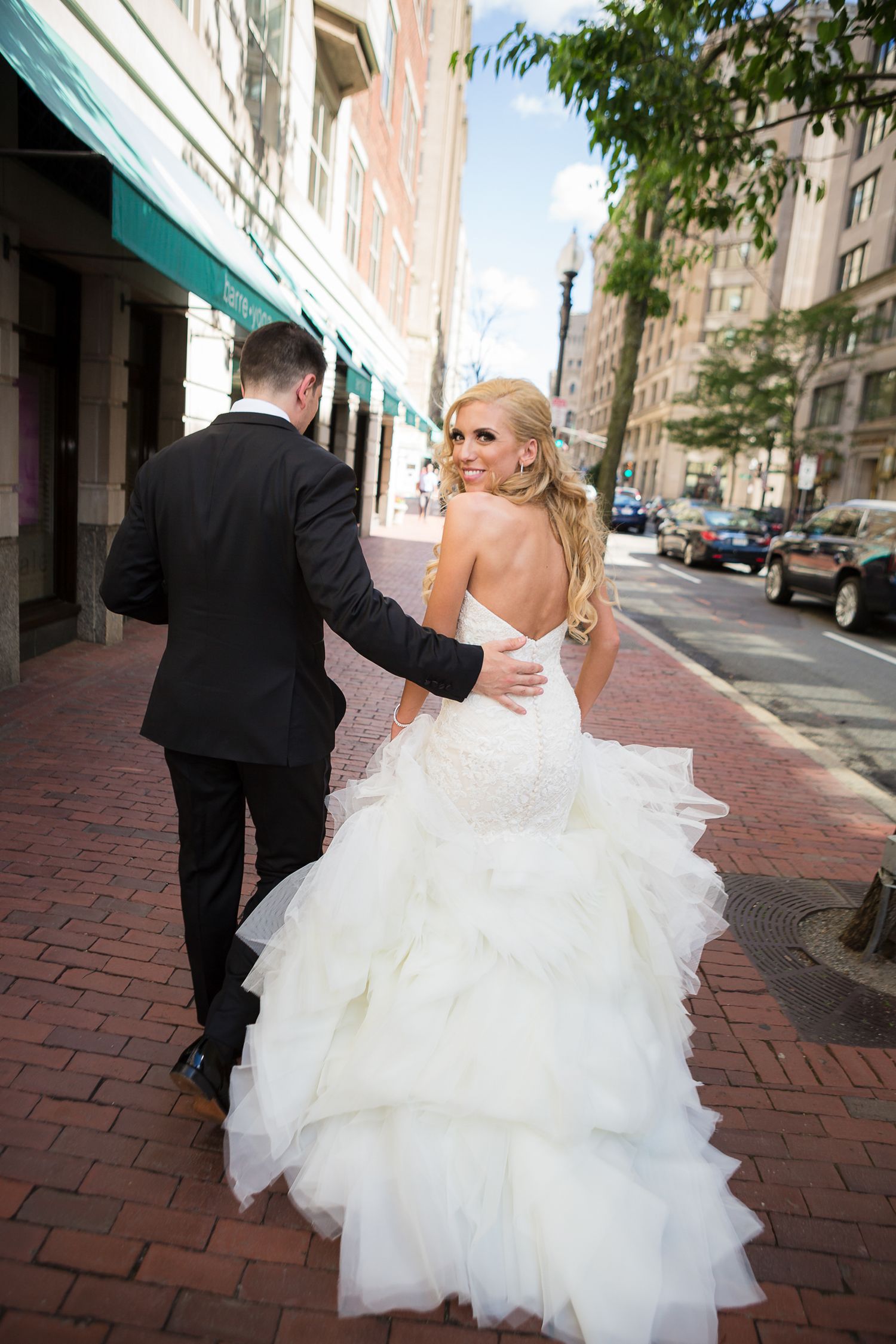 Bride in flowing white dress walks with groom on brick sidewalk in urban setting.