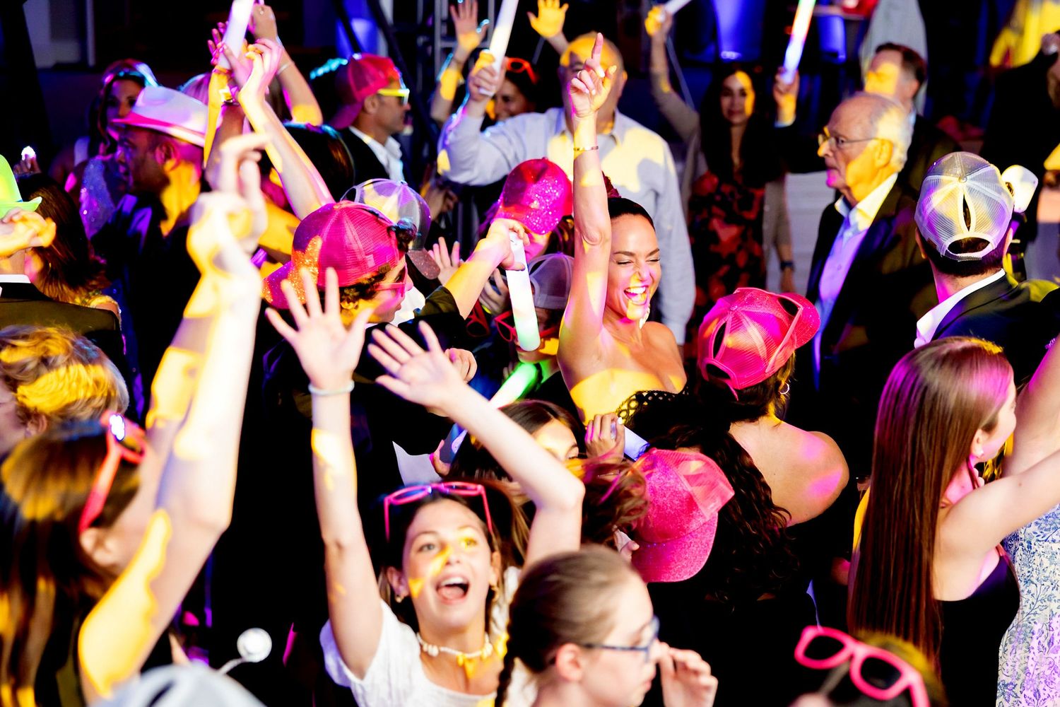 Energetic crowd wearing party accessories and dancing under colorful event lighting.