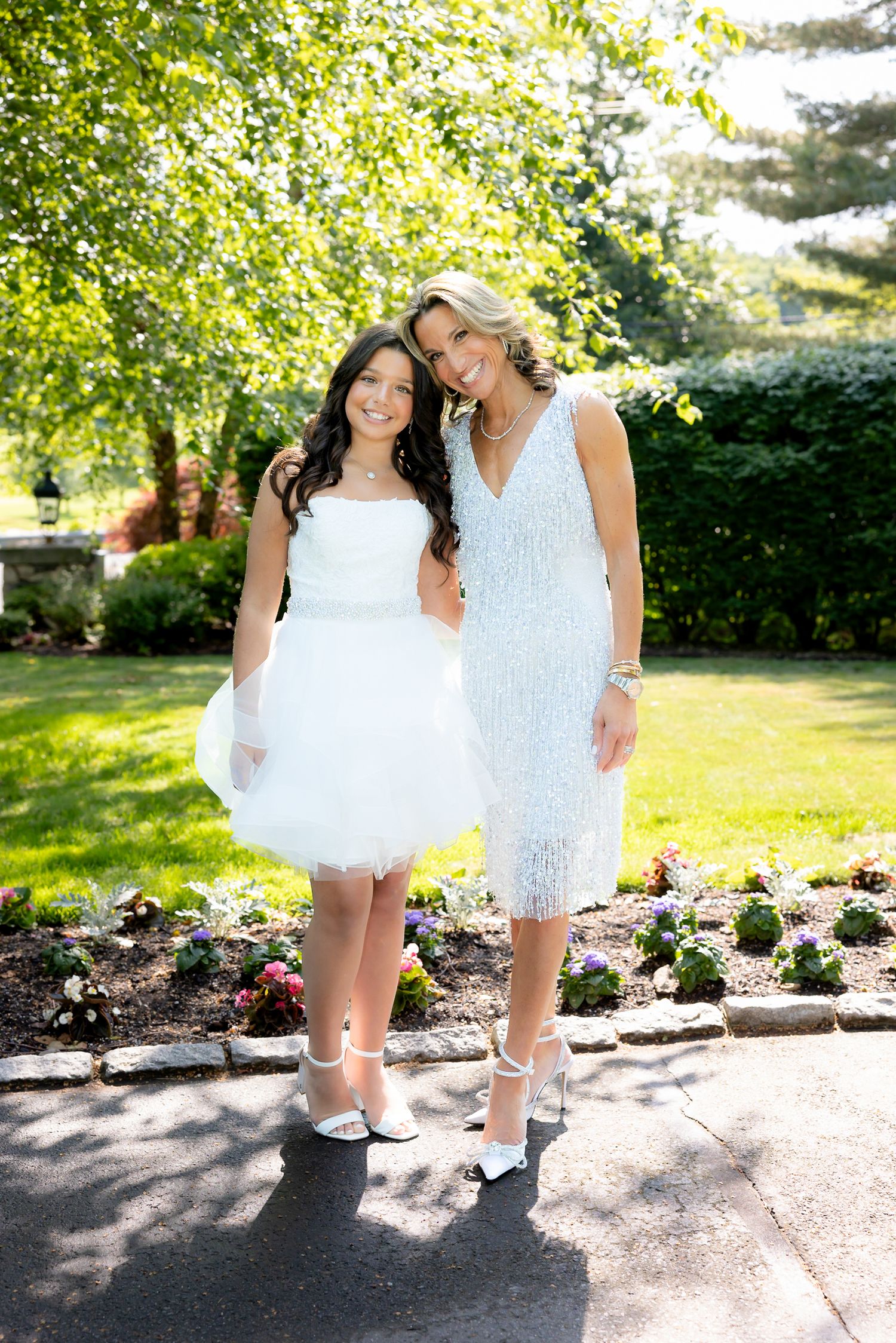Two people in white and lace dresses pose together on a garden path.