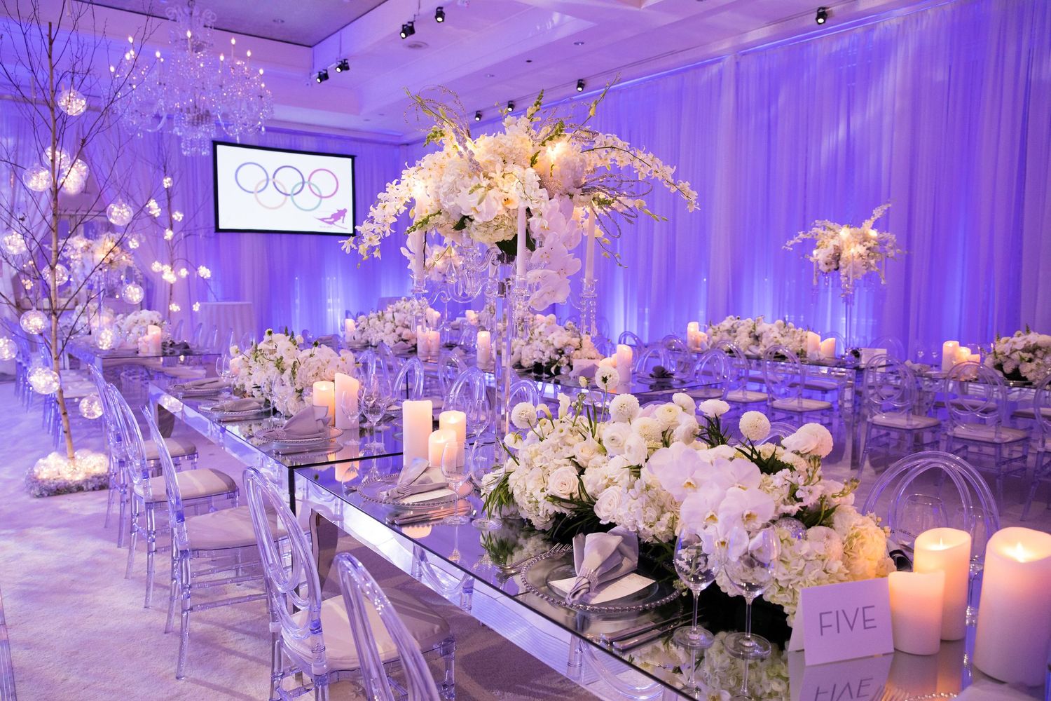 Luxurious white floral centerpieces and candles on mirrored tables under purple lighting.