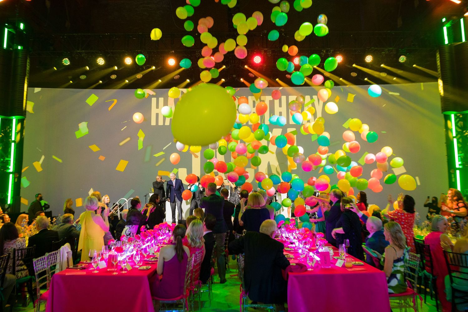Vibrant party scene with colorful balloon drop over pink-lit dining tables.