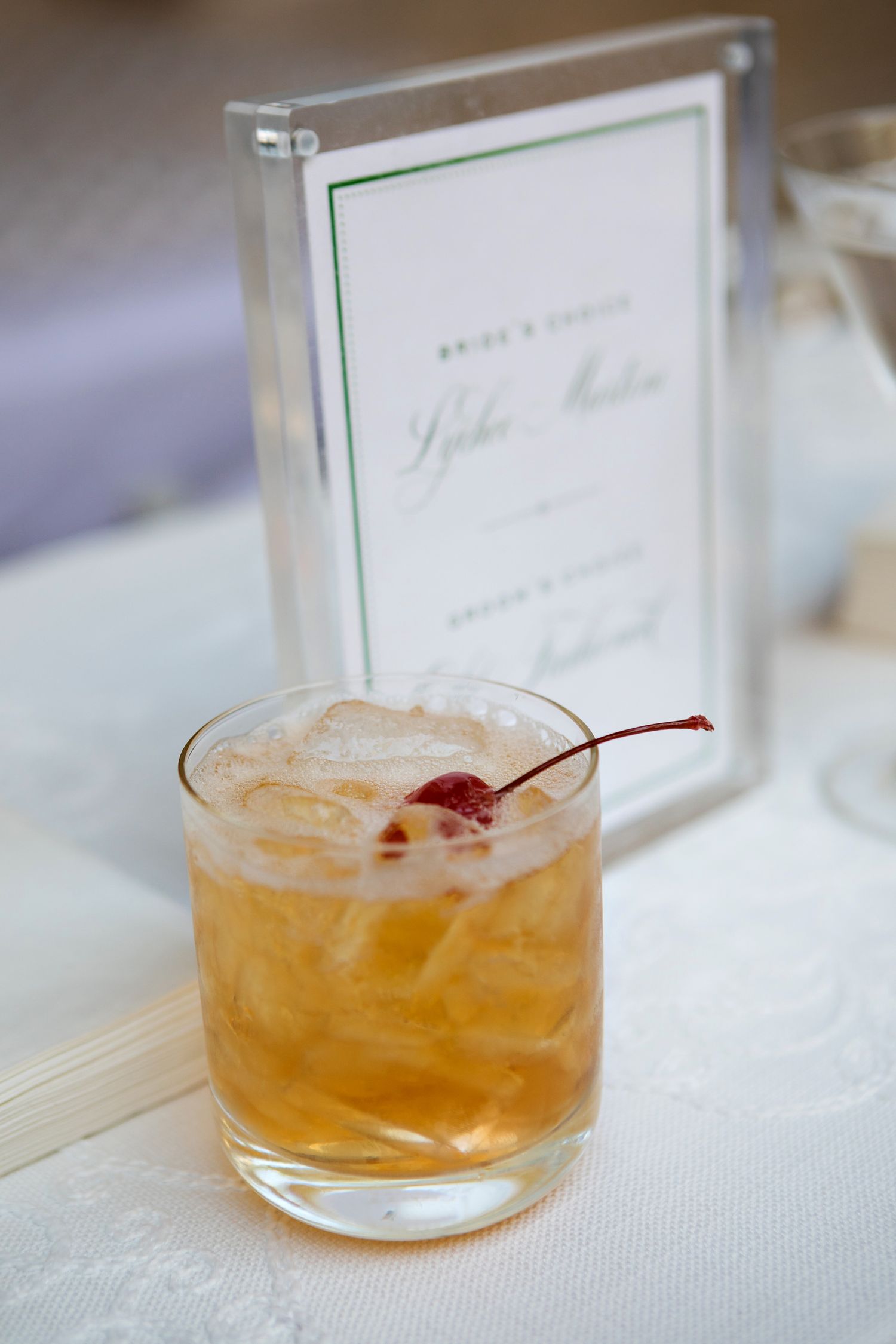 Classic cocktail in rocks glass garnished with a maraschino cherry next to a framed menu.