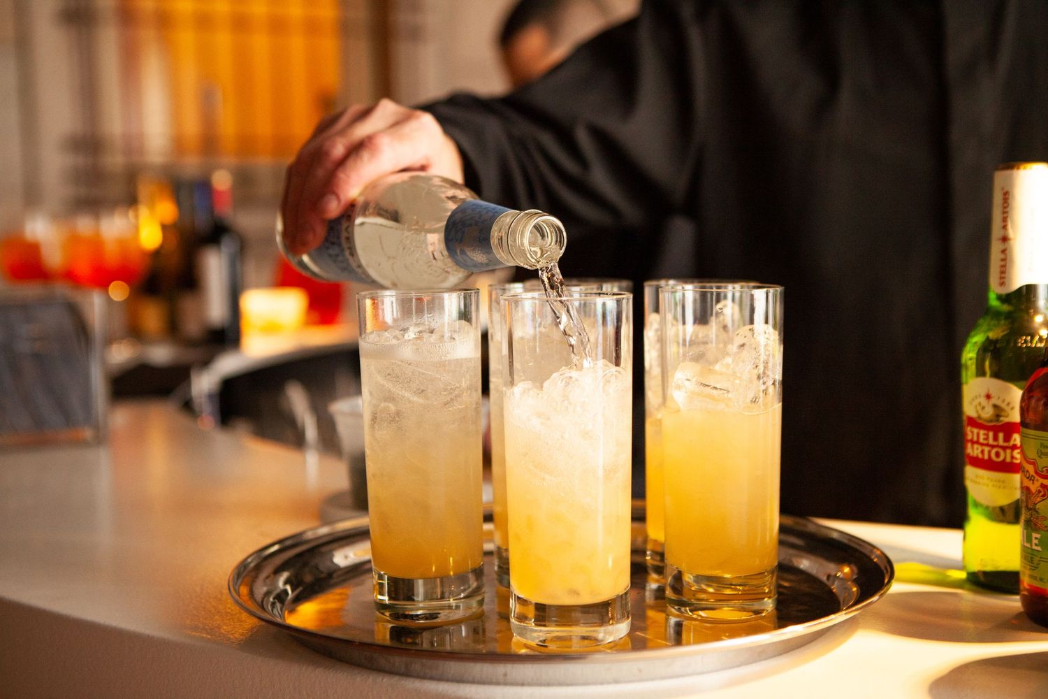 Bartender pouring golden cocktails into tall glasses on a serving tray at an upscale bar.