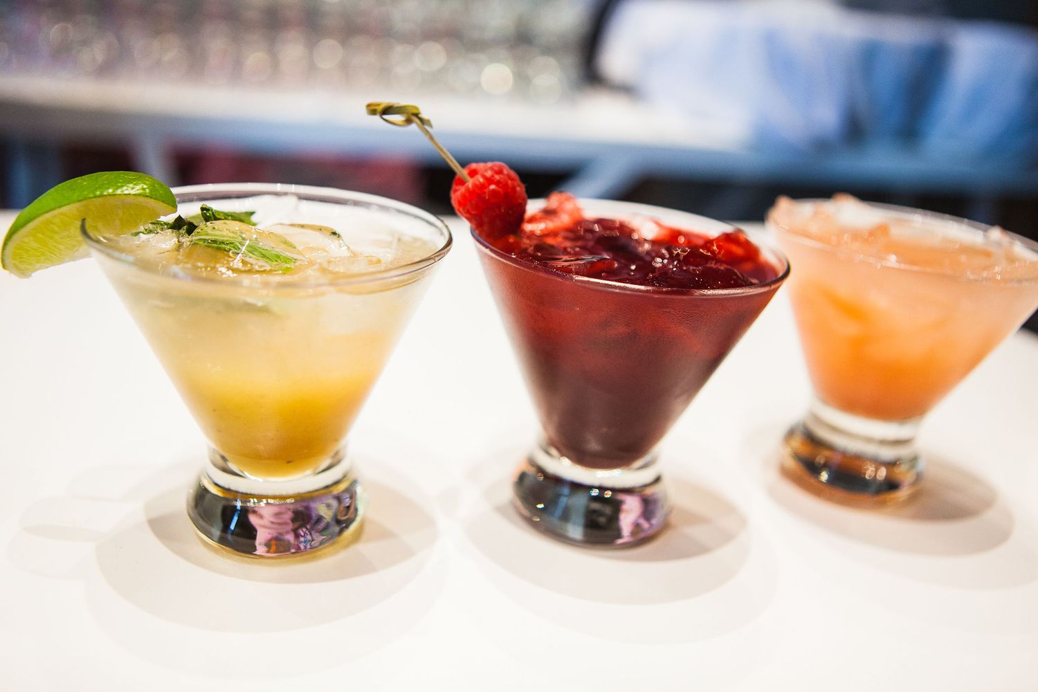 Three colorful cocktails in martini glasses garnished with fruit on a bar counter.