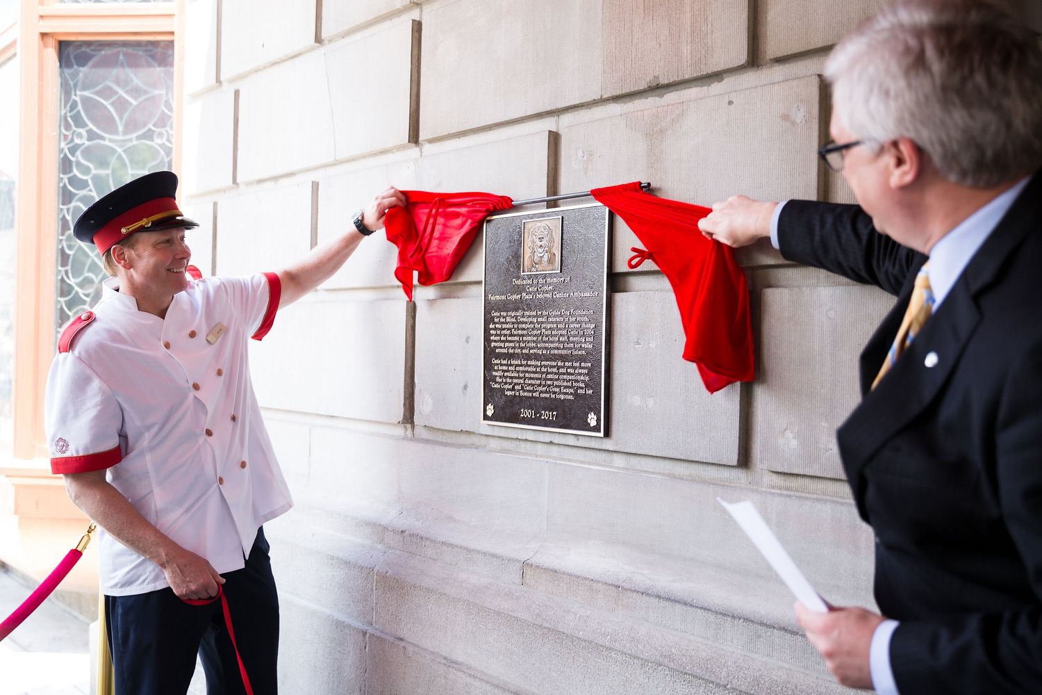 Two people unveiling a commemorative plaque mounted on a white brick wall during a ceremony.