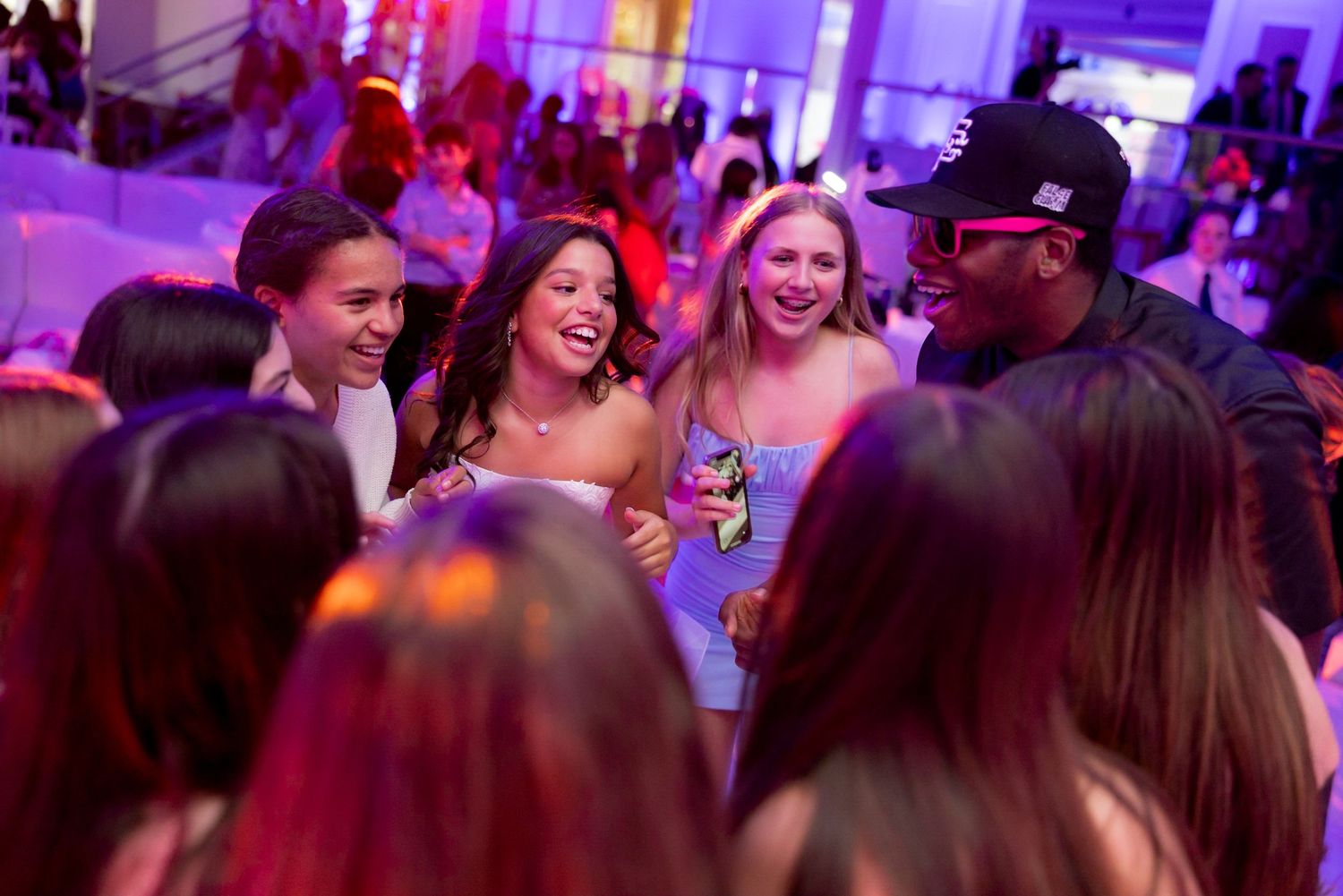 Group of party guests laughing and socializing under pink and purple ambient lighting.