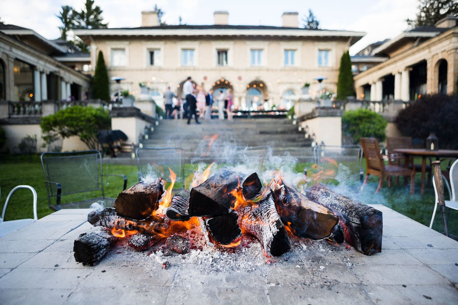 Outdoor firepit with burning logs creates ambiance in front of an elegant estate building.