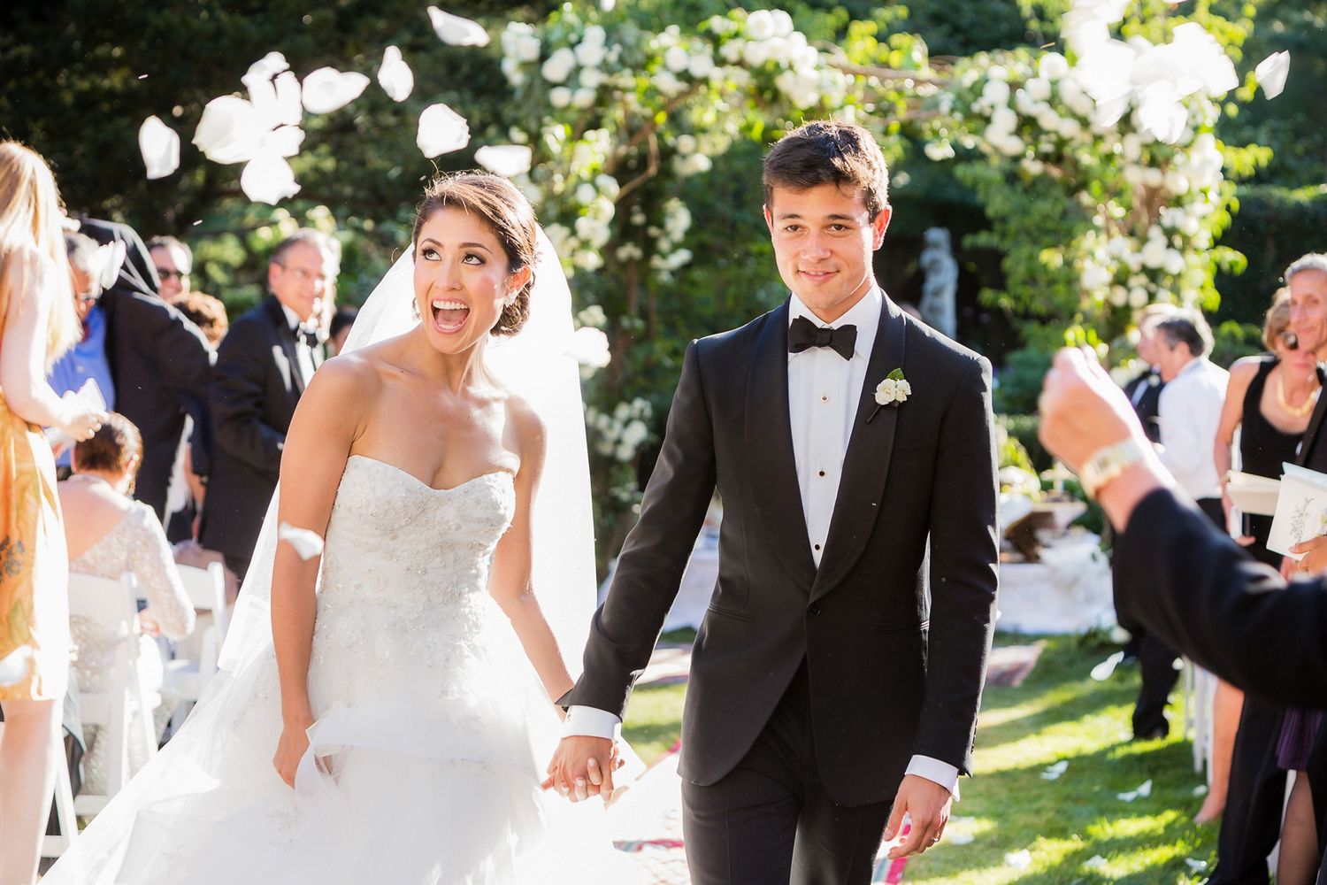 Wedding party members dance and celebrate outdoors in a sunlit garden setting during a joyful reception.