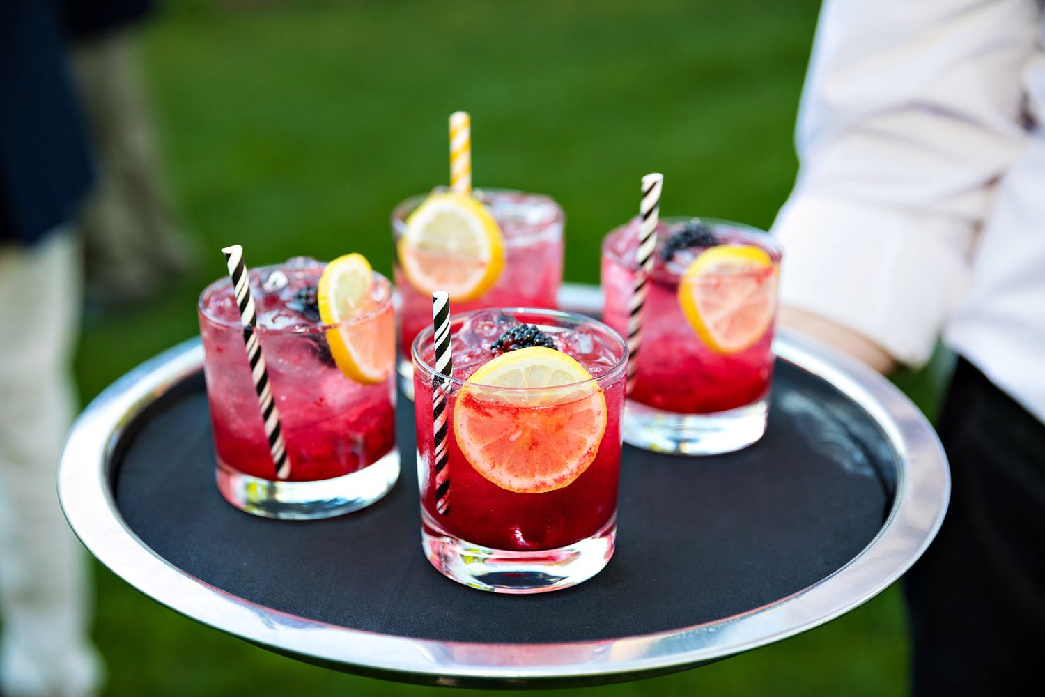 Red cocktails garnished with citrus and striped straws served on a black serving tray outdoors.