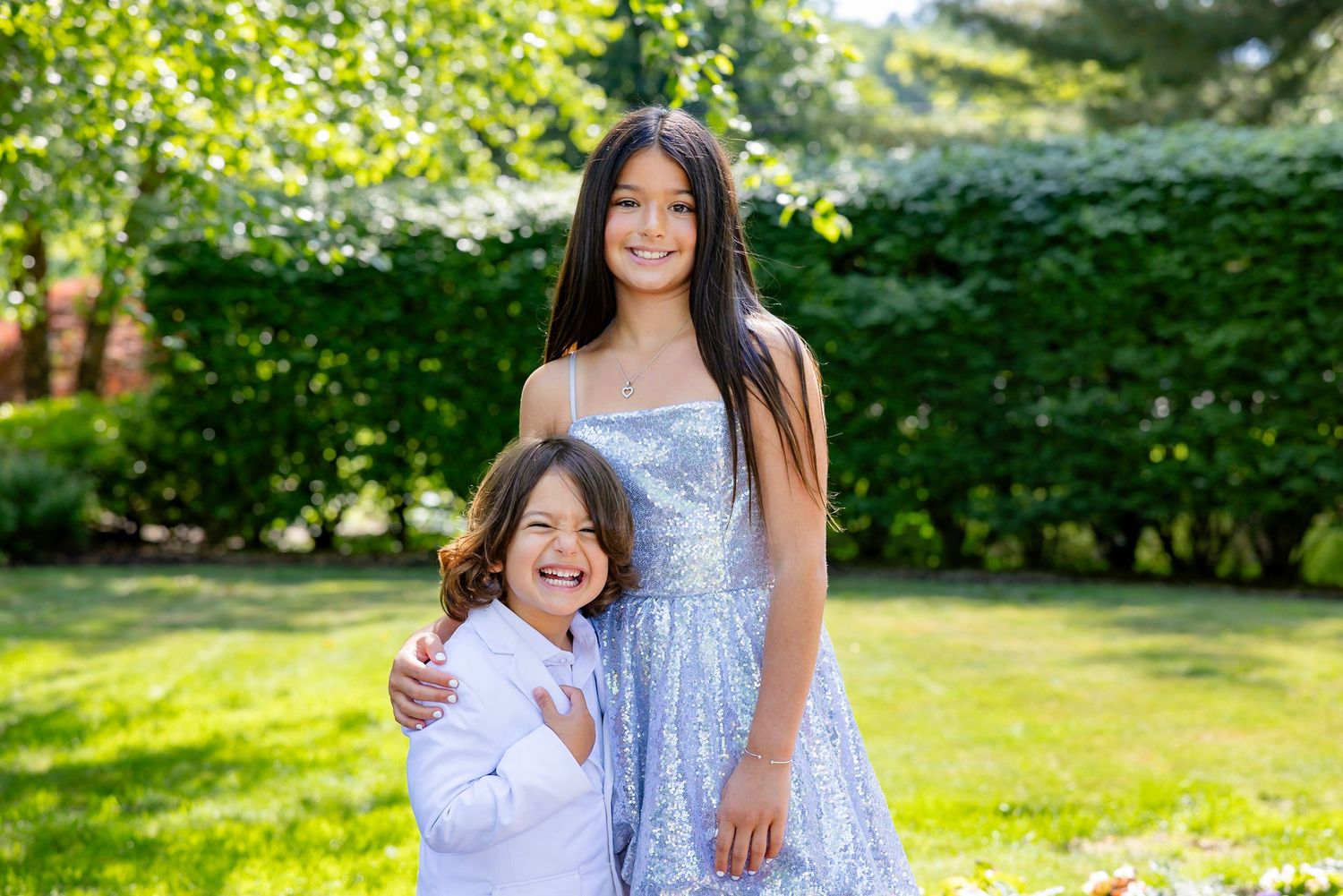 Two people wearing formal attire embrace and smile in a sunny garden setting.