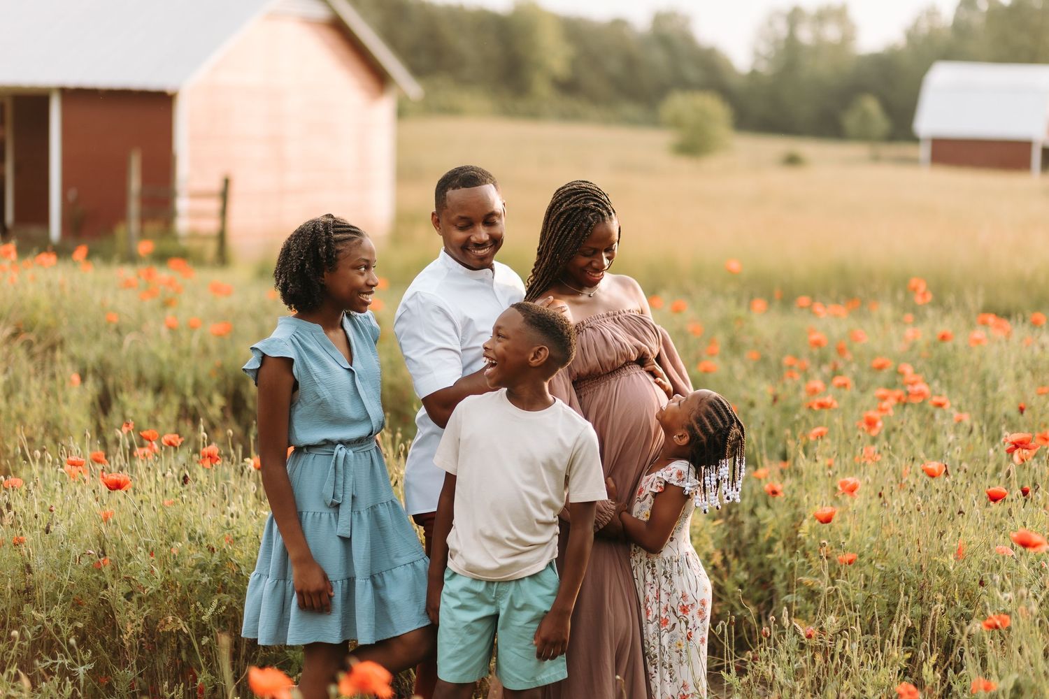 Family of five posing in a vibrant poppy field near a red barn, sharing a joyful moment together.