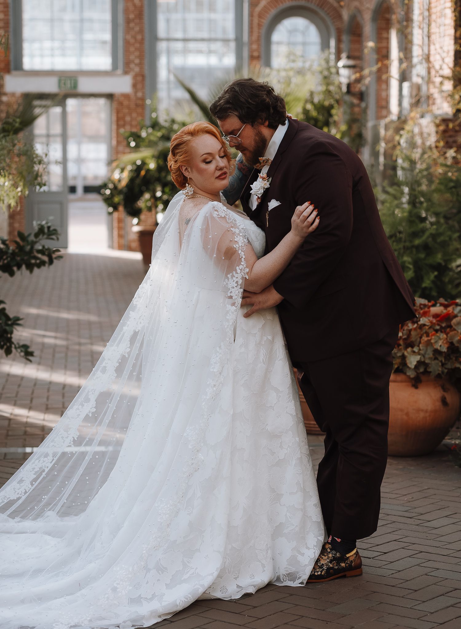 Romantic wedding photo of couple embracing in a conservatory with trailing lace veil and floral decor.