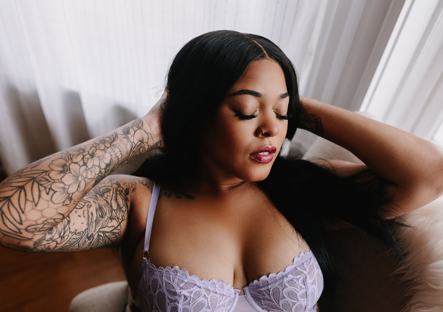 Woman in lavender lingerie poses seductively with eyes closed and visible arm tattoo.