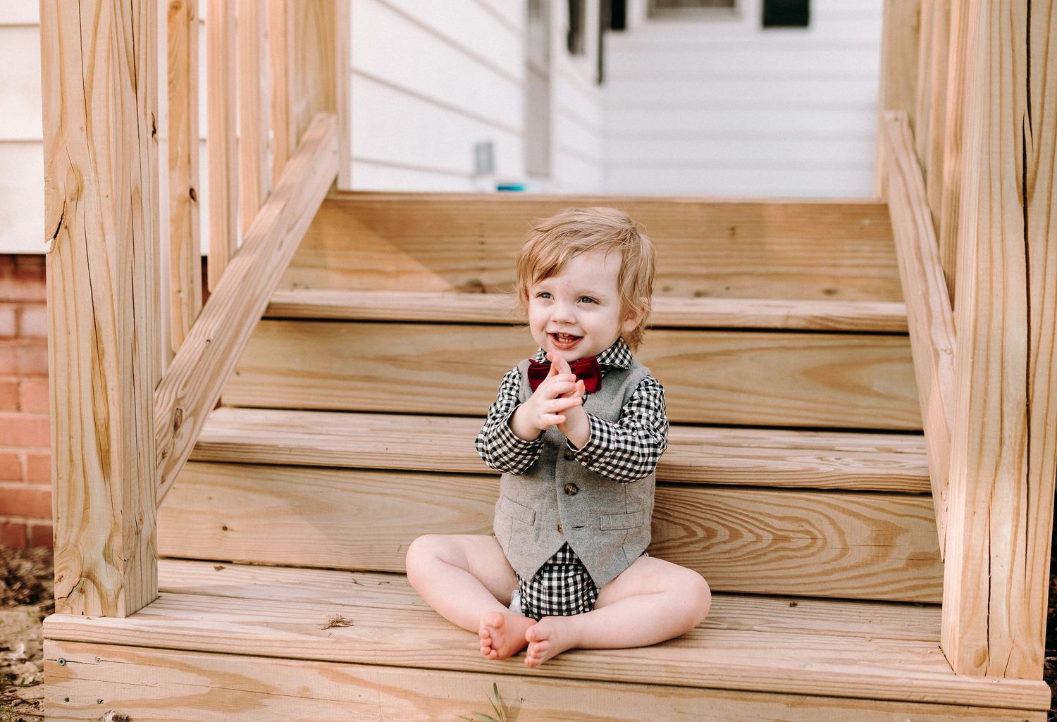 Small child sitting on wooden steps eating something while wearing checkered outfit.