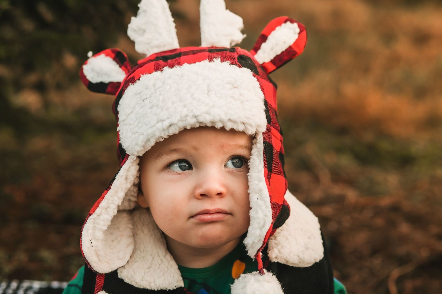 Adorable toddler wearing a festive red plaid reindeer hat with antlers and fluffy white trim outdoors.