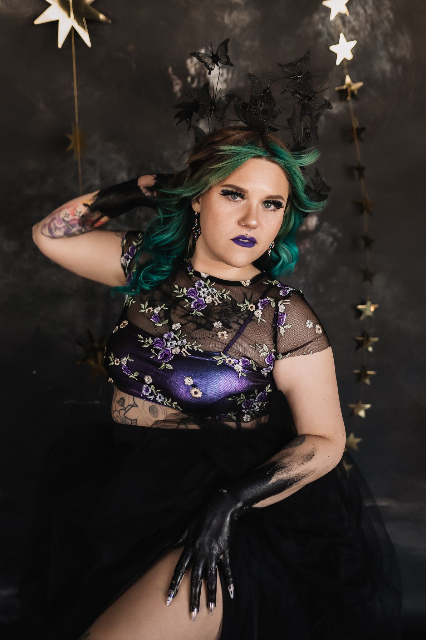 Woman with green hair in black sheer floral top, dark gloves, and star decorations, styled in gothic Halloween costume.