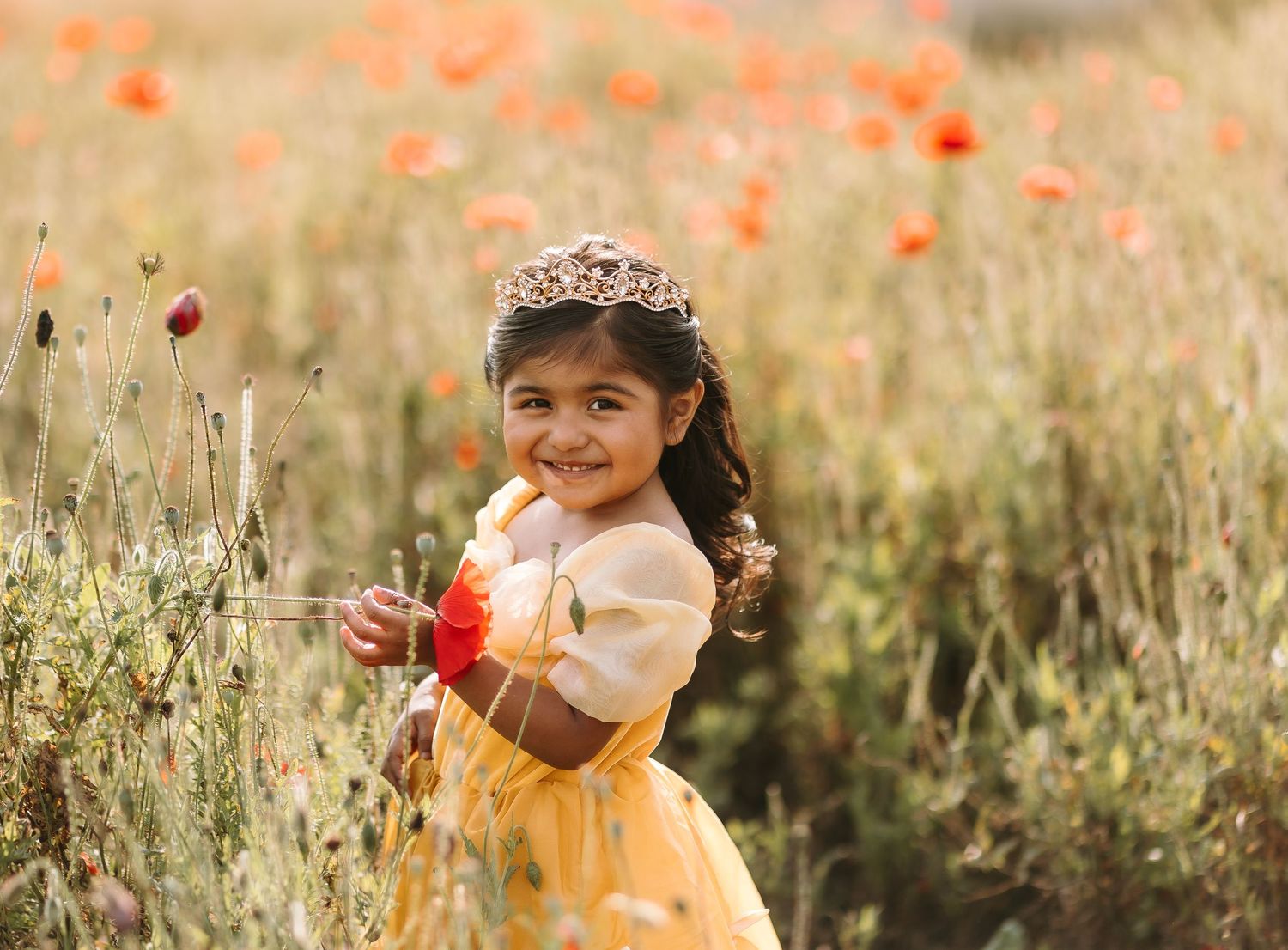 Smiling little girl in yellow princess dress and tiara holding flowers in a vibrant red poppy field.