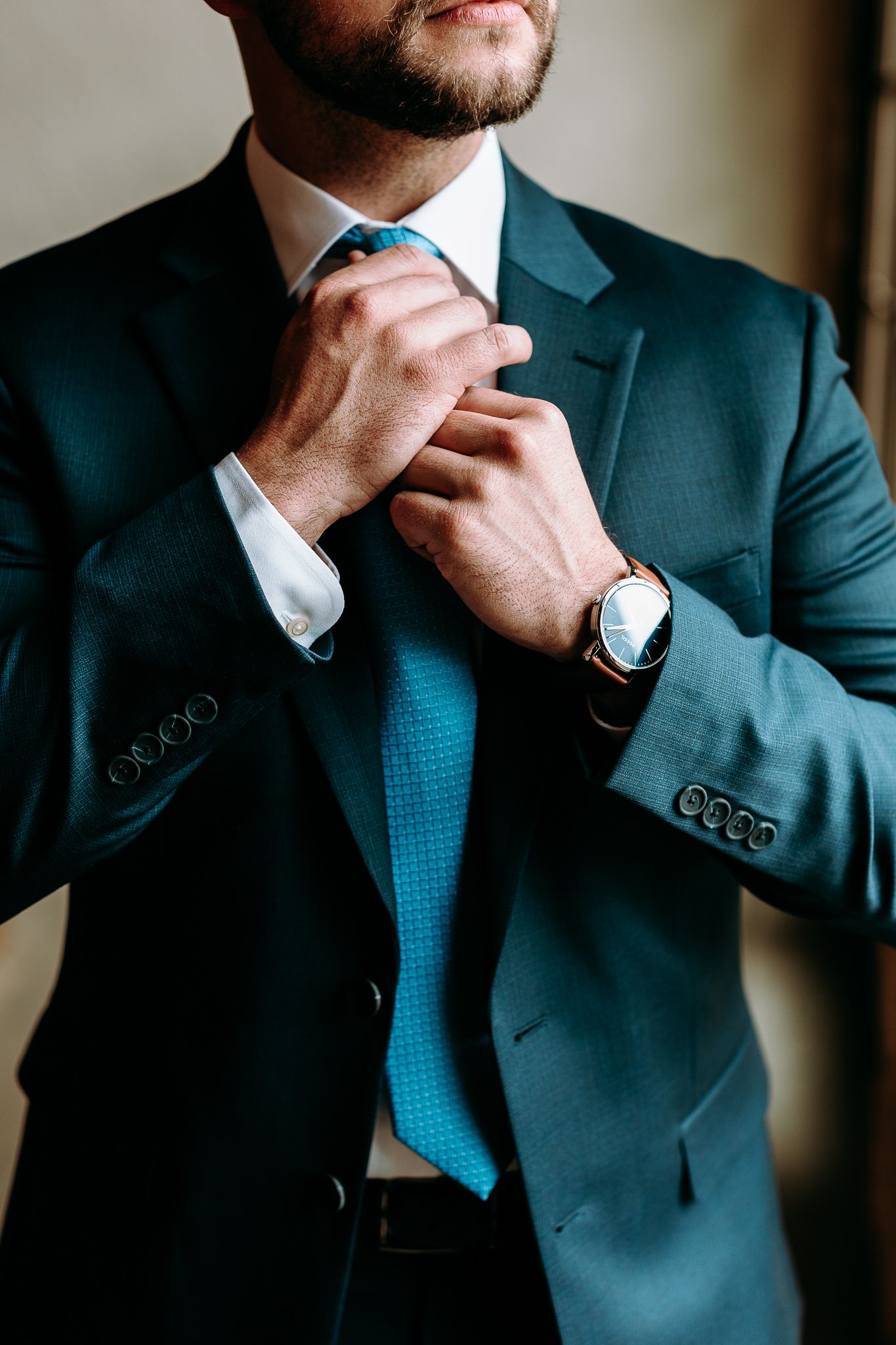 Close-up of man adjusting teal tie wearing navy suit and wristwatch.