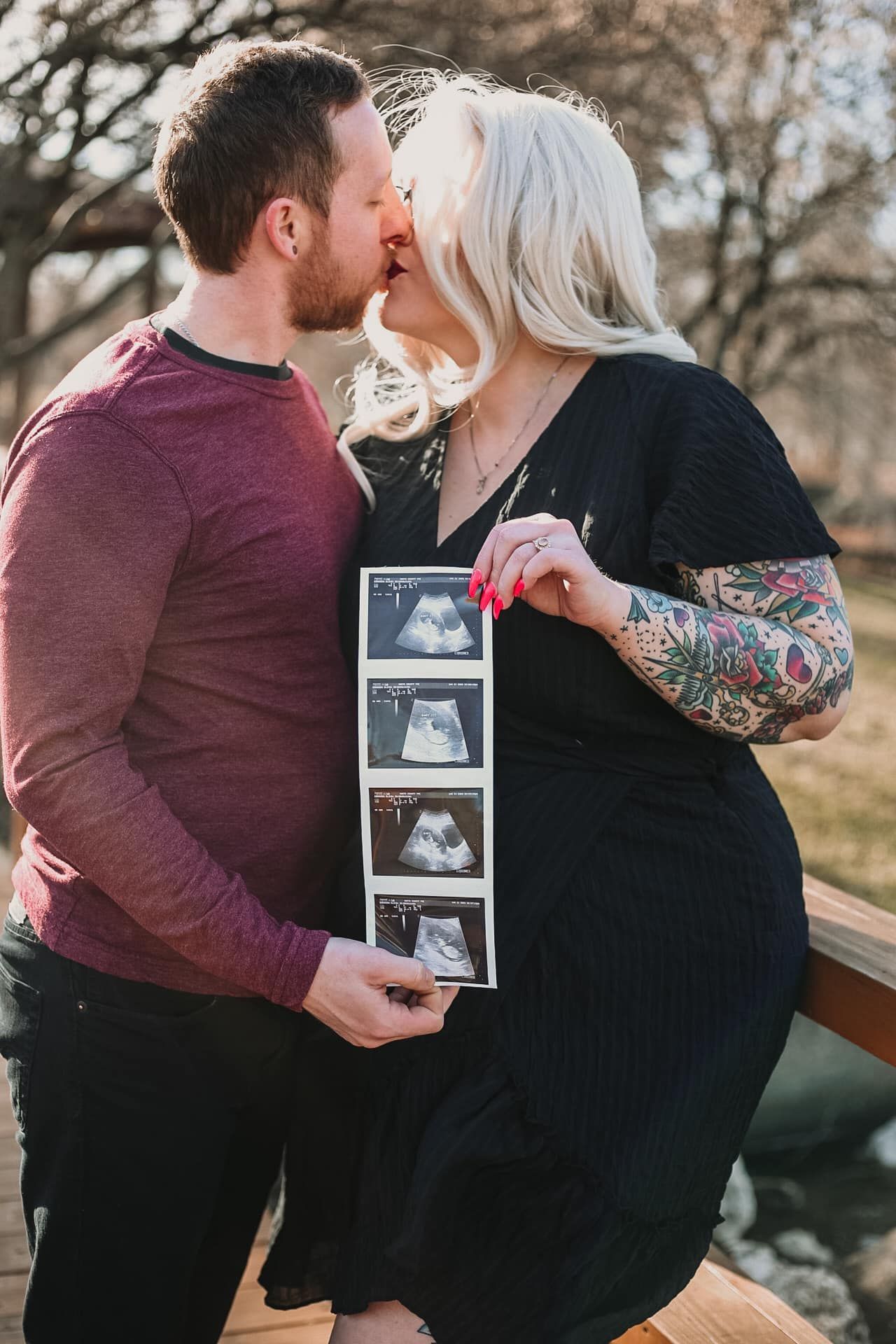 A couple shares an intimate moment while holding ultrasound photos outdoors.